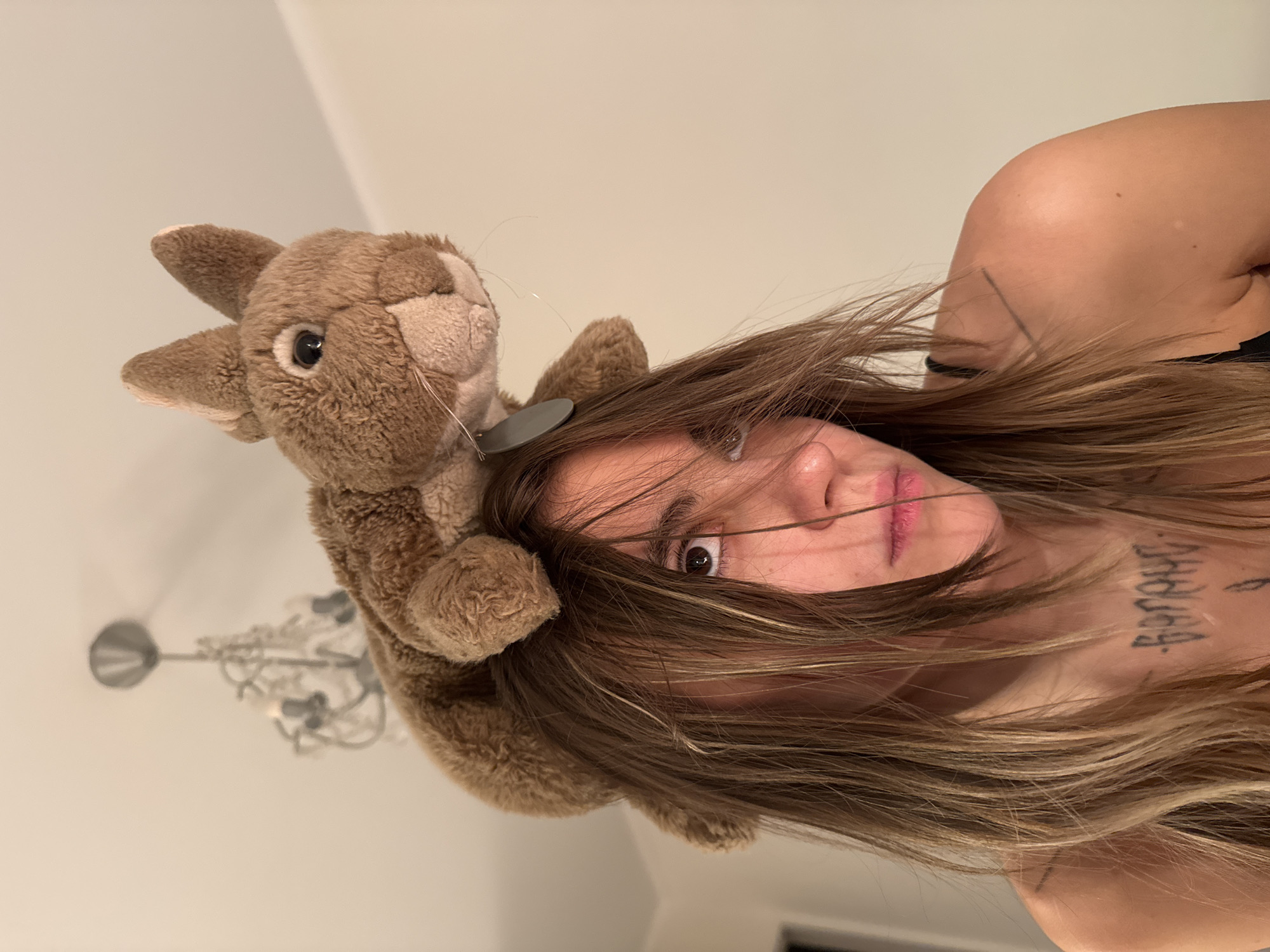 A person with long, wavy, brown hair is looking at the camera. A brown stuffed rabbit toy is resting on top of their head. The person is wearing a black tank top, and has tattoos on their chest and shoulder. The background is a plain, light-colored wall with a chandelier visible at the top left. The scene appears to be indoors. There is no indication of the location being Moscow, Russia.