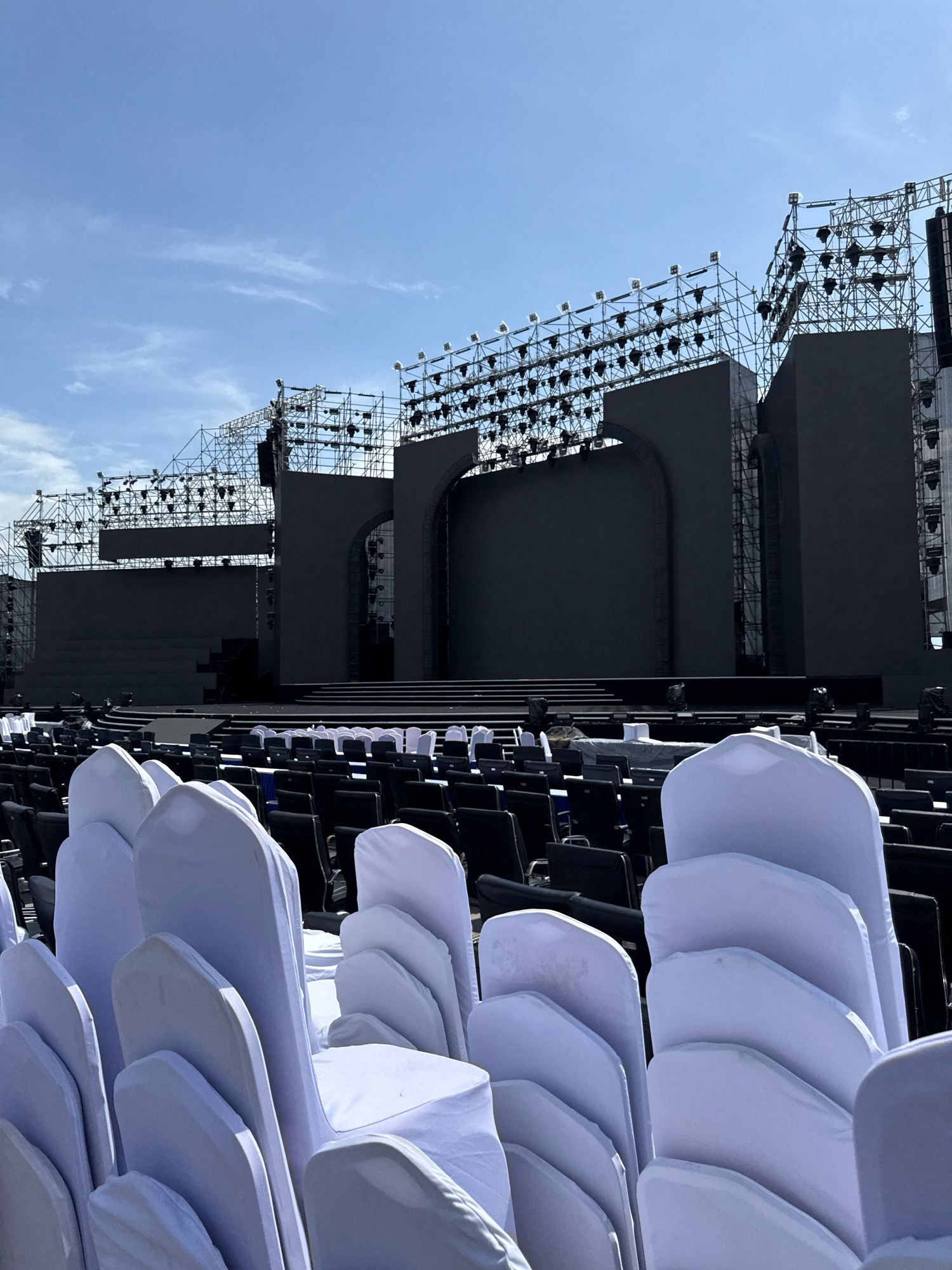 An outdoor stage setup for an event is shown in Nha Trang, Vietnam. The stage features a large, dark-colored screen and a complex scaffolding structure overhead, fitted with numerous lights. In the foreground, rows of chairs are arranged, with many in the front covered in white fabric. The sky above is clear blue with a few wispy clouds. The overall scene suggests preparation for a large-scale performance or ceremony.