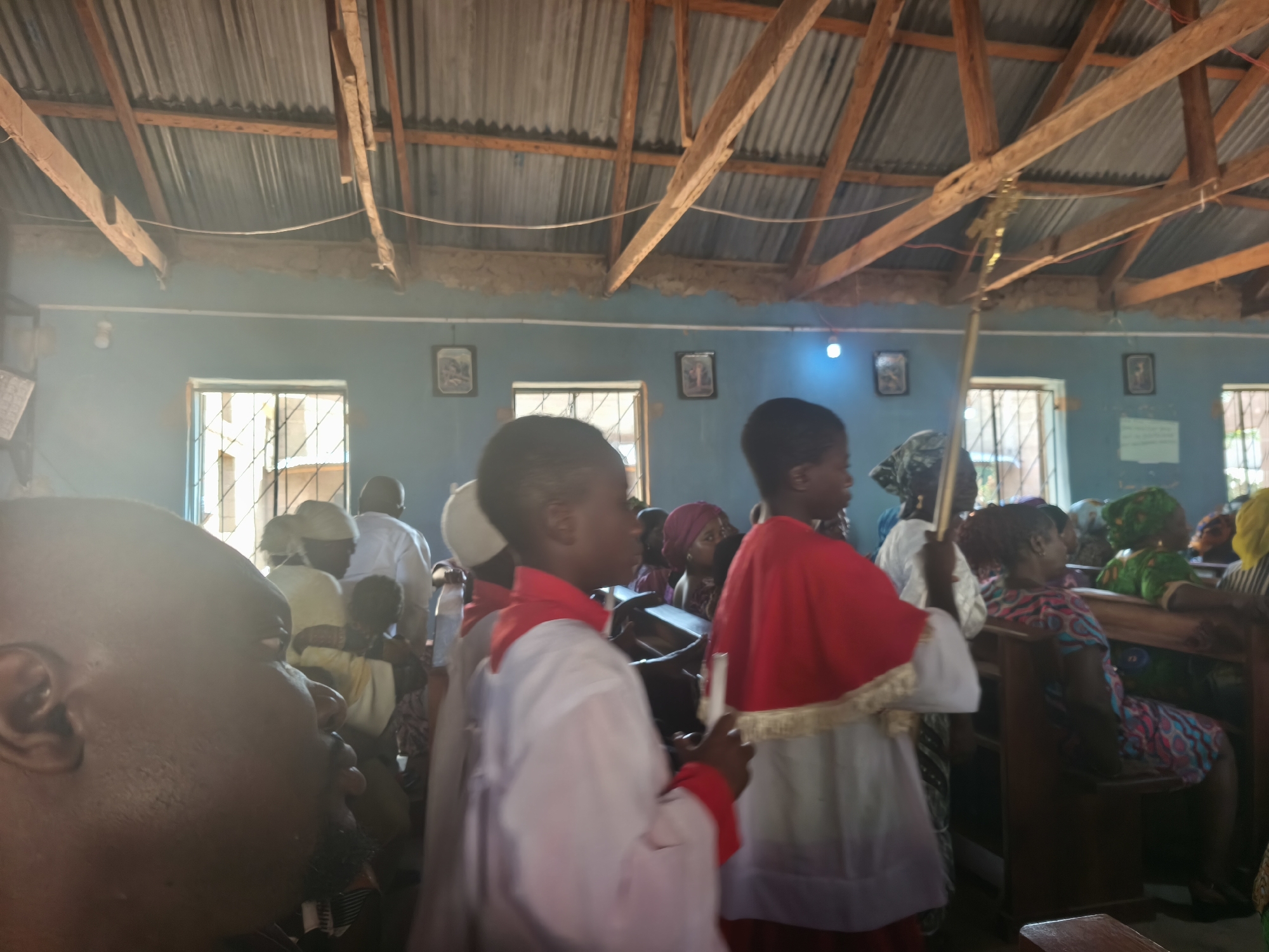 **Setting:** Interior of a church in Nabatum, Nigeria, with a corrugated metal roof supported by wooden beams.
**People:** Primarily African people. Many individuals dressed in white robes with red capes are visible, seemingly participants in a religious procession or ceremony. Some people sit on benches. Other individuals are visible wearing headscarves and patterned clothing.
**Objects:** Wooden benches, windows with bars.
**Environment:** Bright light, interior of a church.
**Action:** Religious ceremony.