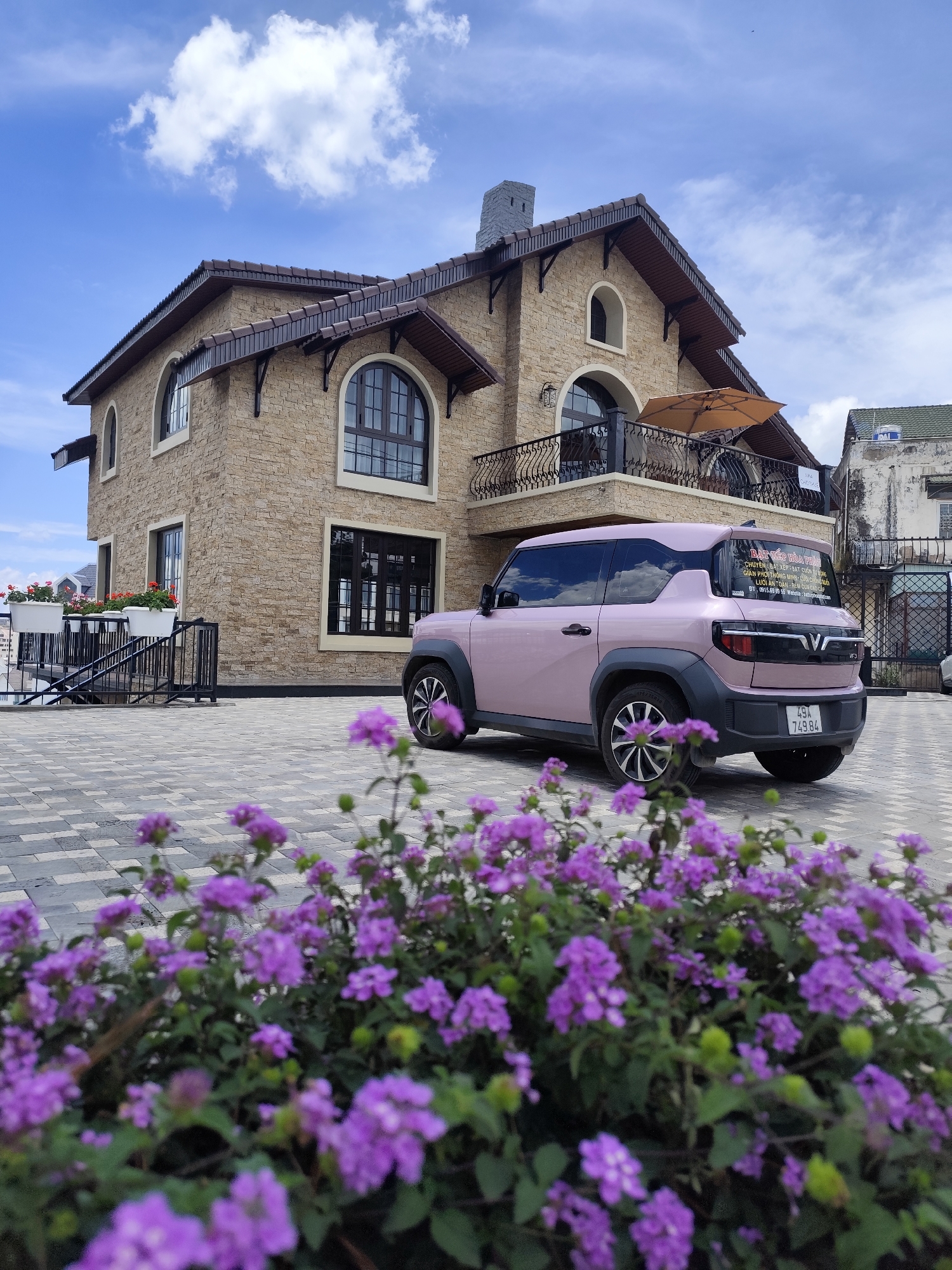 The image presents a scene in Xuân Trường - Đà Lạt, Vietnam. The focal point is a light pink VinFast VF3 electric SUV parked in front of a two-story building with a stone facade. The building features arched windows with dark frames, a balcony with an umbrella, and a chimney on the roof. A black wrought iron staircase leads to an upper level, and flower boxes adorn the railing. In the foreground, a lush bush of purple flowers partially obscures the view. The car has a sticker with Vietnamese text on the rear window, and the license plate number is 49A-749.84. The parking area is paved with interlocking gray stones. The sky is blue with scattered white clouds. Another building is visible adjacent to the main structure.