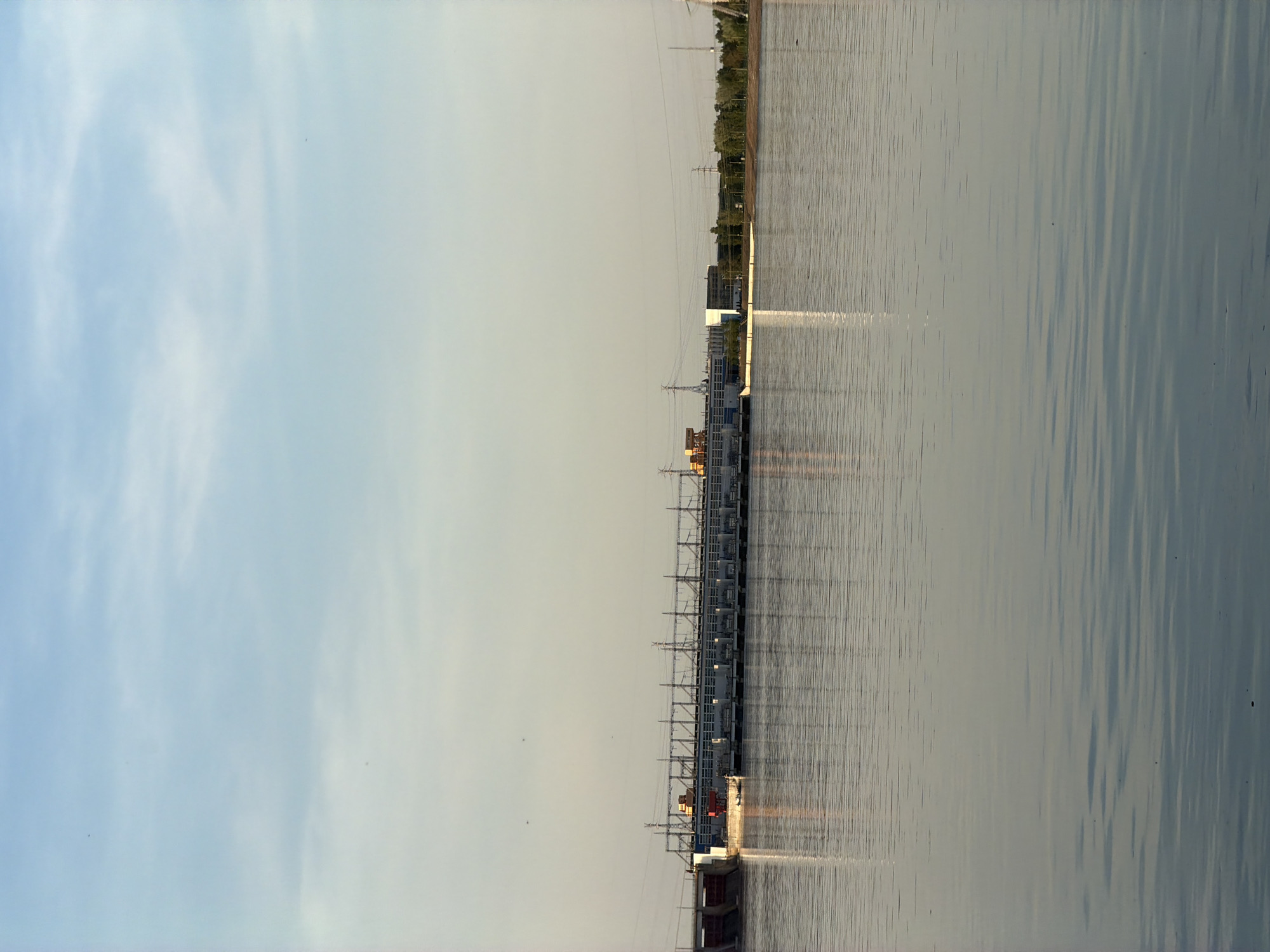 The image depicts a water power plant in Noviy, Russia. The plant stretches across the frame, with its infrastructure and equipment visible. Water fills the foreground and stretches into the distance. The sky is a pale blue with some wisps of clouds. Power lines extend from the plant towards the background, where some trees can be seen. The lighting indicates it might be early morning or late afternoon. There are no people visible in the shot.