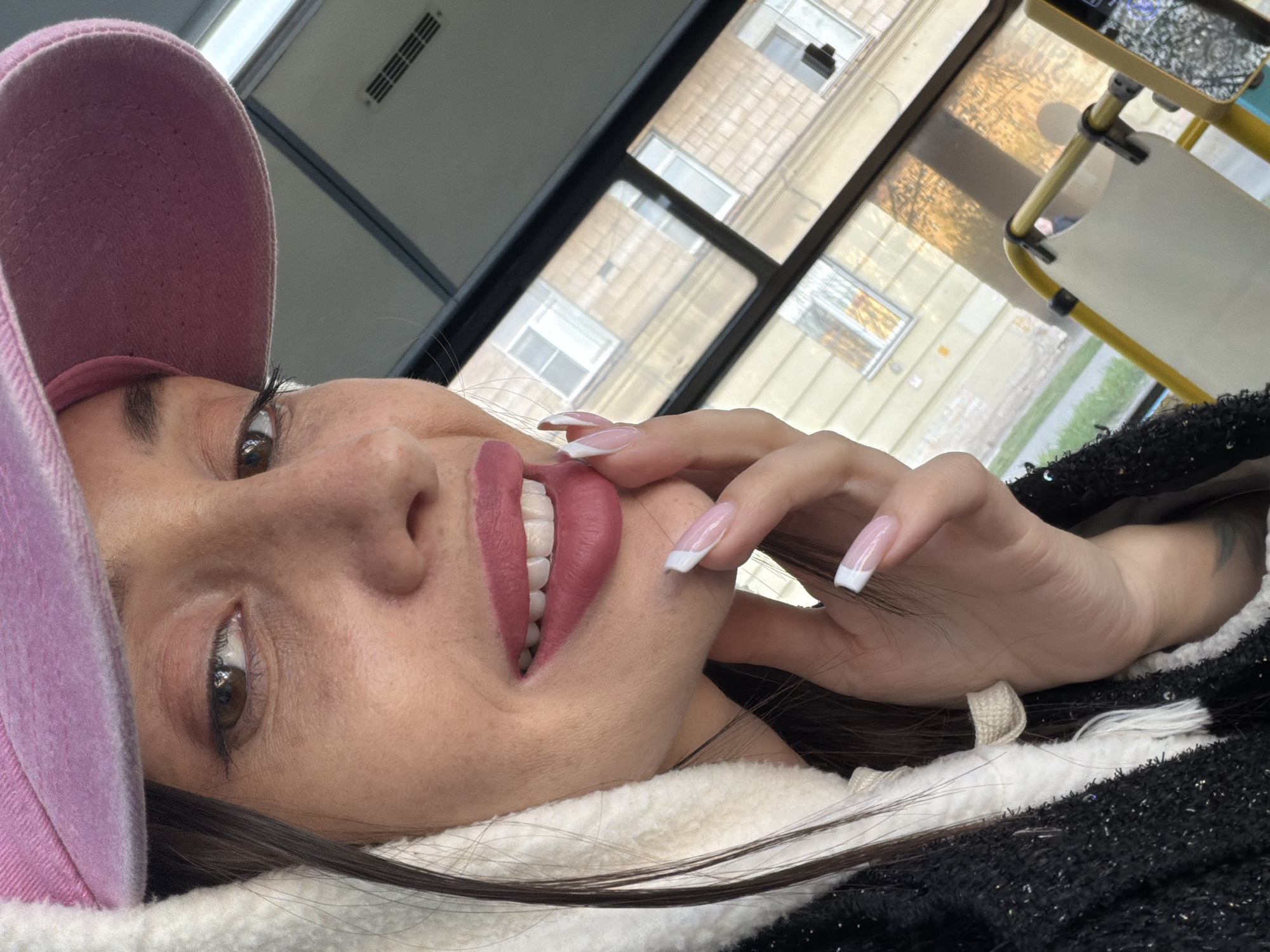 A young woman with light skin, wearing a pink baseball cap and a white-trimmed black jacket, is posing for a selfie. She has long, dark hair and a hand near her face, showcasing white-tipped nails. The background is a window showing buildings with apartments and a street with greenery. The scene is in the city of Saint Petersburg, Russia, based on the buildings' style.