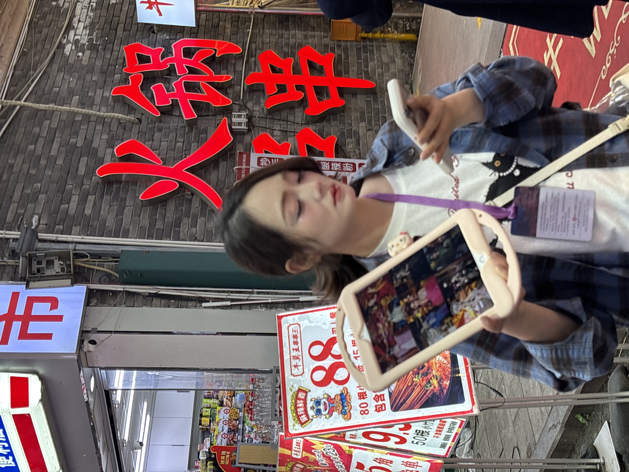 Here is a detailed description of the image:
A young child is holding a tablet in front of a storefront in the Yuzhong District of China. The child is facing the camera, but their gaze is directed downwards towards the screen of the tablet. They are wearing a white T-shirt with a black cat design, covered by a blue plaid button-down shirt. A lanyard with an ID badge is visible around their neck.
Behind the child, there is a storefront with several signs in Chinese. A large, red sign with Chinese characters is mounted on a dark brick wall. Below this, there's an illuminated white sign with red lettering. Further down, a smaller sign advertises "牛莫王串串王" (Niu Mo Wang Chuan Chuan Wang) with the price "88". The storefront appears to be filled with various products, visible through the glass entrance.
The street setting is urban, with a sidewalk and other pedestrian barriers visible. Overall, the image captures a moment of the child's interaction with technology in the context of a commercial environment in a Chinese city.