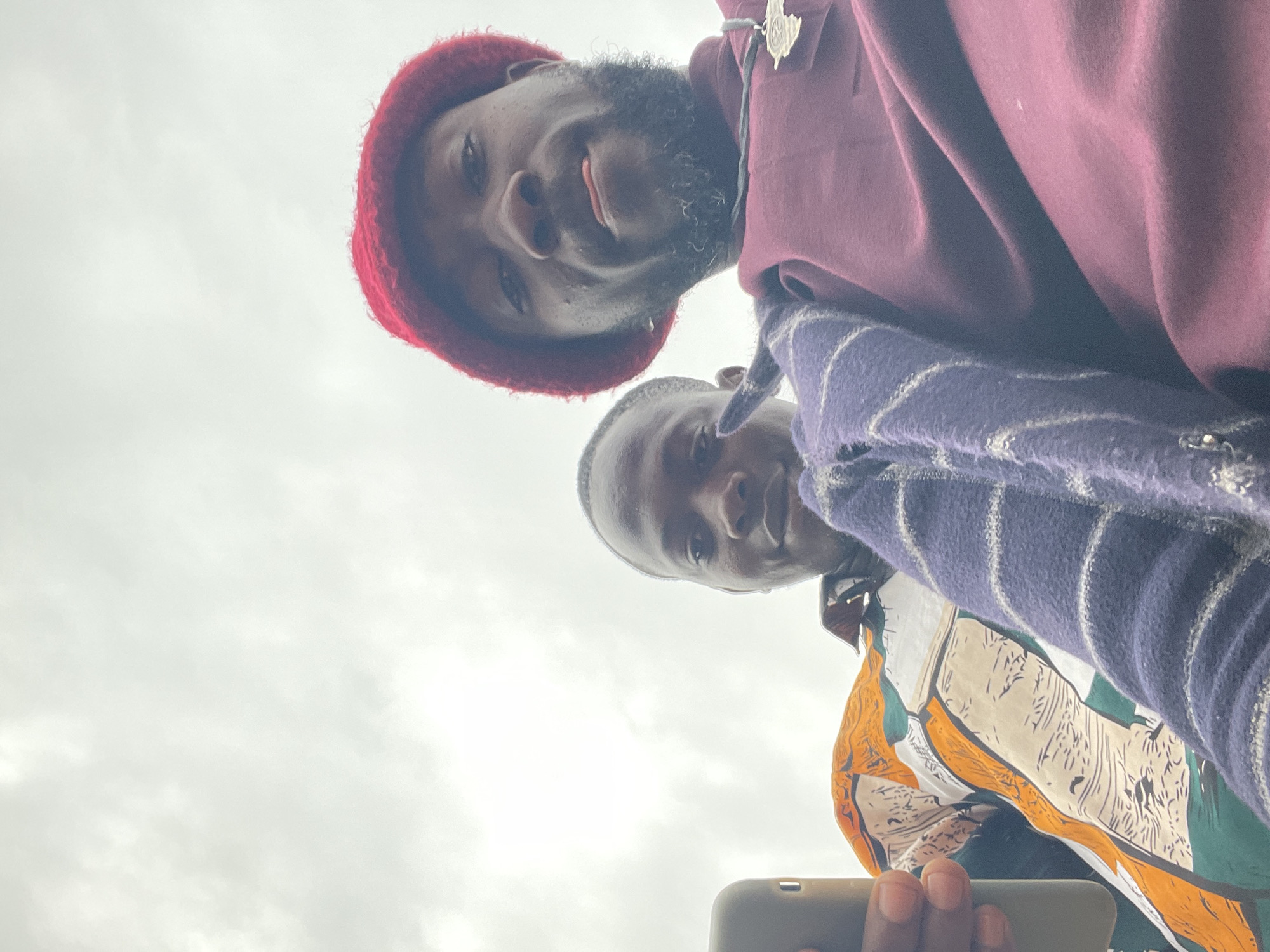 Two Black males are captured in a low-angle, selfie-style shot against a cloudy sky, suggesting an outdoor setting possibly in the city of Jos, Nigeria. The male in the foreground is wearing a red beanie and a burgundy shirt, with a silver pendant hanging from his neck. He has a beard and appears to be sticking his tongue out slightly, looking directly at the camera. The other male, positioned slightly behind and to the left, is wearing a colorful shirt with an abstract pattern of orange, white, green, and black. He is holding a mobile phone in his left hand, which is visible at the left edge of the frame. A portion of a purple and white striped garment is draped across the chest area of the individuals. The overall impression is a candid moment, possibly shared between friends.