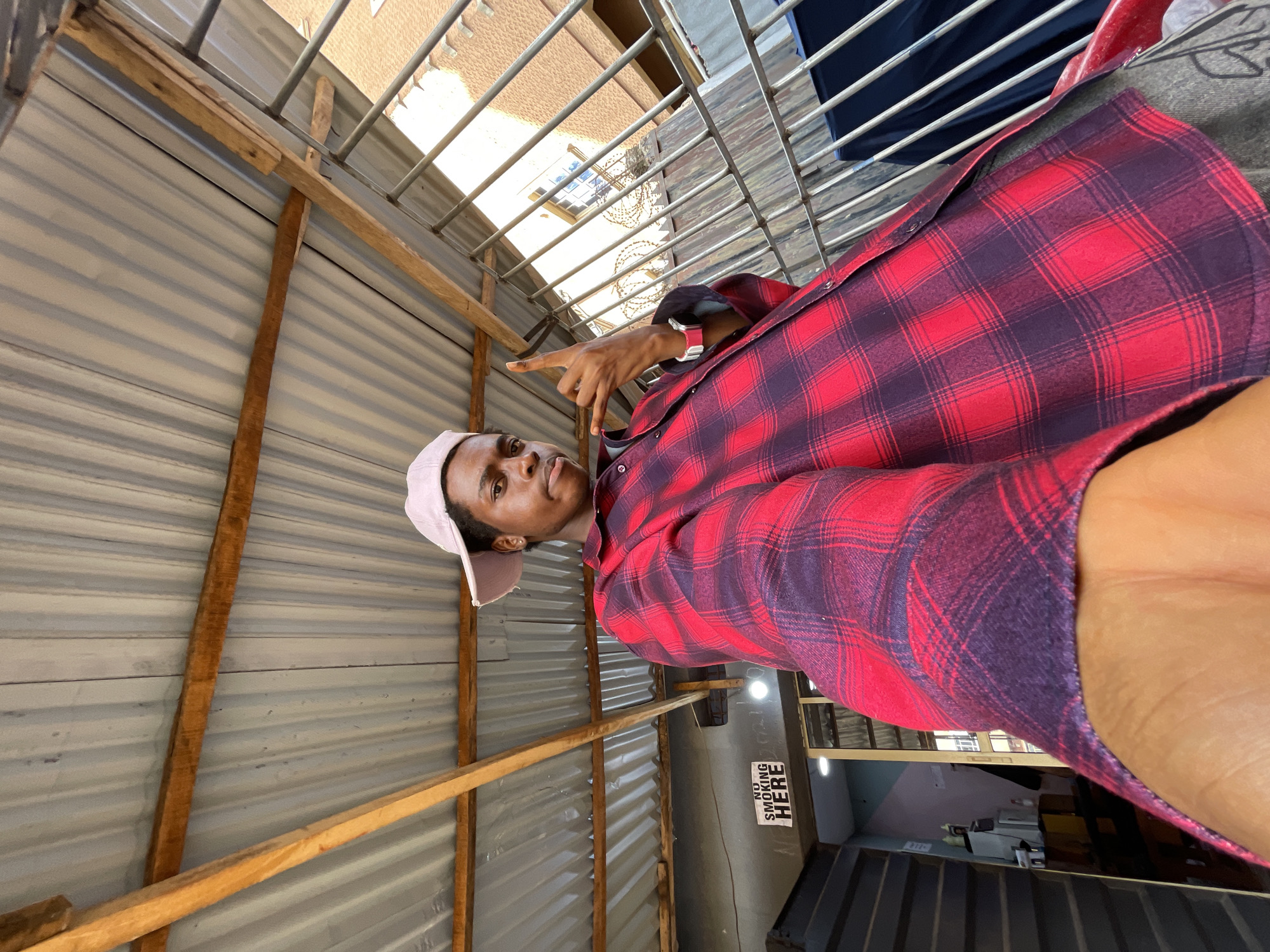 The image shows a man in a red and purple plaid shirt and a pink cap, gesturing with his left hand. He appears to be in a covered area with wooden beams and corrugated metal roofing. Beyond the beams, there is a metal grating and a view of a building with windows and barbed wire. A sign reading "NO SMOKING HERE" is visible on a wall. Based on the architecture and the sign, the image might be taken in a commercial or industrial area. The image's composition suggests it was taken as a selfie or at a low angle. Based on the architecture and the signage, it is plausible that the photo was taken in Onitsha, Nigeria.