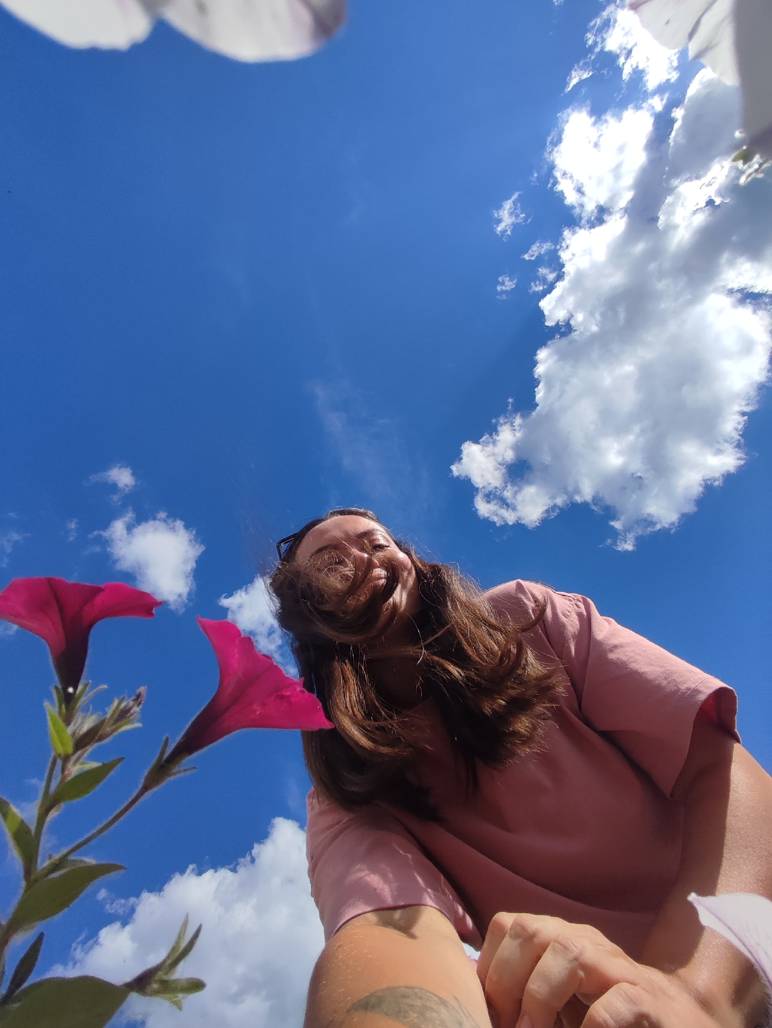 The media file is a close-up, low-angle shot of a person outdoors on a sunny day. The person, with long brown hair, is looking up at the camera and smiling broadly, with their hair blowing across their face. They are wearing a light pink t-shirt. Two vibrant pink petunia flowers are in the foreground, with stems and leaves visible. The background is a bright blue sky filled with fluffy white clouds, suggesting a clear and pleasant weather. The sun is shining, casting light on the clouds and creating a bright atmosphere. The shot appears to be taken in a garden or park. The overall mood is cheerful and energetic. There is no visible text in the media file. The location context is Chaikovsky, Russia.