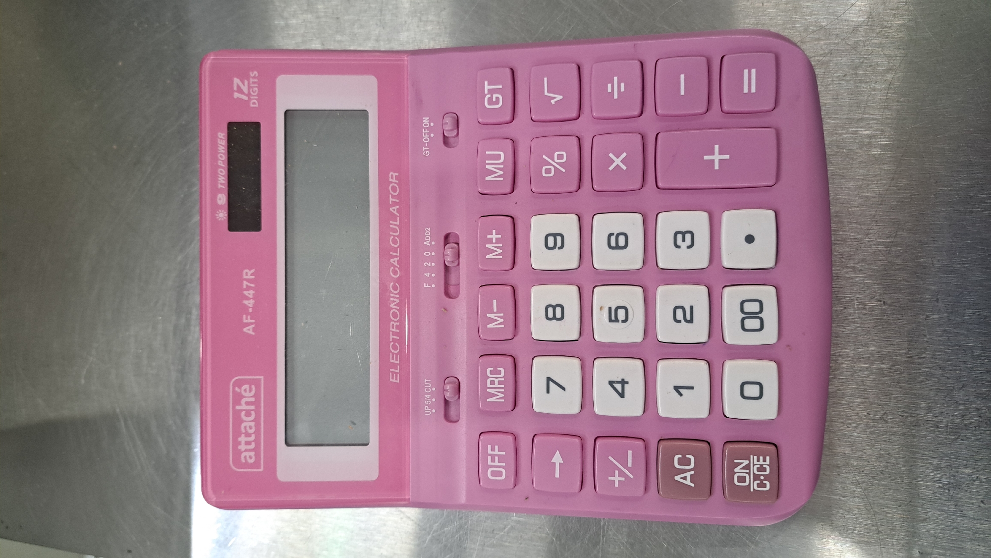 This is a top-down, eye-level shot of a pink electronic calculator, model AF-447R by Attaché. The calculator is placed on a metallic, textured surface that appears to be a table or countertop. The calculator is the sole subject of the image, and no people are visible. The scene is an indoor setting, likely a home or office, with no discernible time of day or weather cues.
The calculator features a large LCD screen on the left side. To the right of the screen, there are various function buttons including "GT", "MU", "M+", "M-", "MRC", "OFF", and "AC". The numerical keypad consists of buttons for digits 0 through 9, a decimal point, and a "00" key. Mathematical operation buttons like "+", "-", "x", "÷", "%", and "√" are also present. There are also controls labeled "GT-OFF ON" and a slider with options "F 4 2 0 ADD2" and "UP 5/4 CUT". The top row of function keys includes "GT", "√", "÷", and what appears to be an equals sign or pause symbol. The calculator also has a button labeled "ON C-CE". The overall design is functional with clearly marked buttons. The metallic surface shows a brushed or scratched texture, suggesting it might be stainless steel.