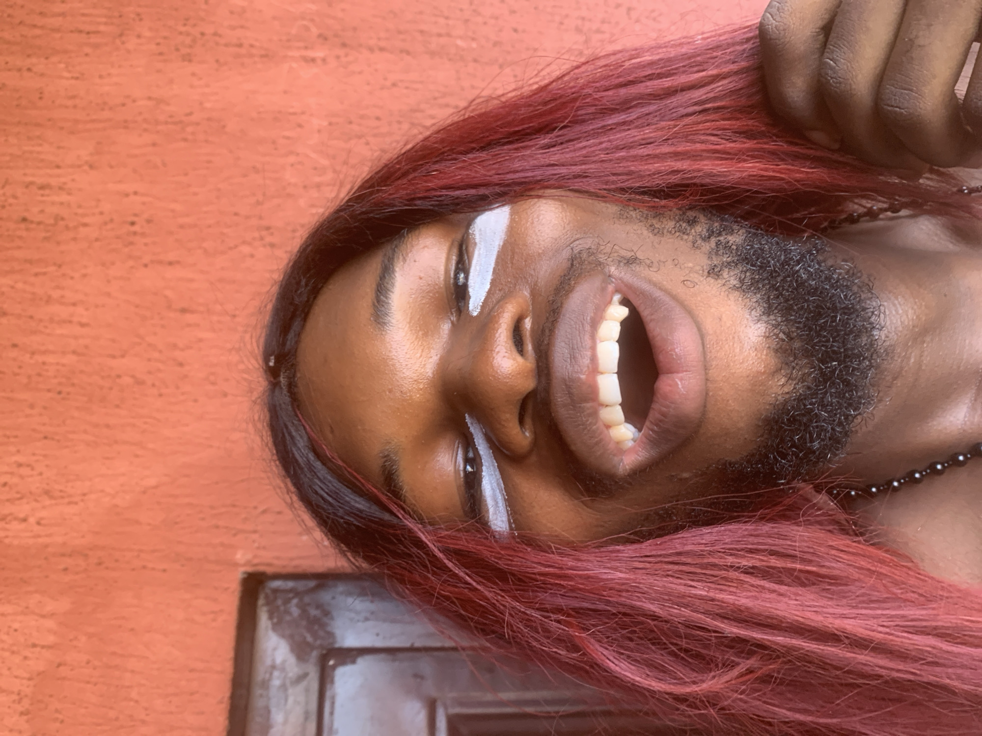A close-up, sideways portrait captures a person with dark skin and a vibrant reddish-burgundy wig or dyed hair, smiling broadly. The individual is positioned against a textured, earthy orange wall, suggesting an outdoor or brightly lit indoor setting in Nkpor, Nigeria. White paint or makeup is applied in a distinct stripe directly under each eye. The person has a neatly trimmed dark beard and is captured mid-laugh or enthusiastic smile, revealing their teeth. Their eyes are looking upwards, conveying a joyful or excited emotion. A hand with dark skin, likely belonging to the subject, is visible at the top of the frame, near their hair. A black beaded necklace is partially visible around their neck. In the lower right corner, a dark, rectangular element, possibly part of a door frame or wooden structure, is visible. The lighting is bright and natural, indicating it is daytime. There is no visible text in the image.