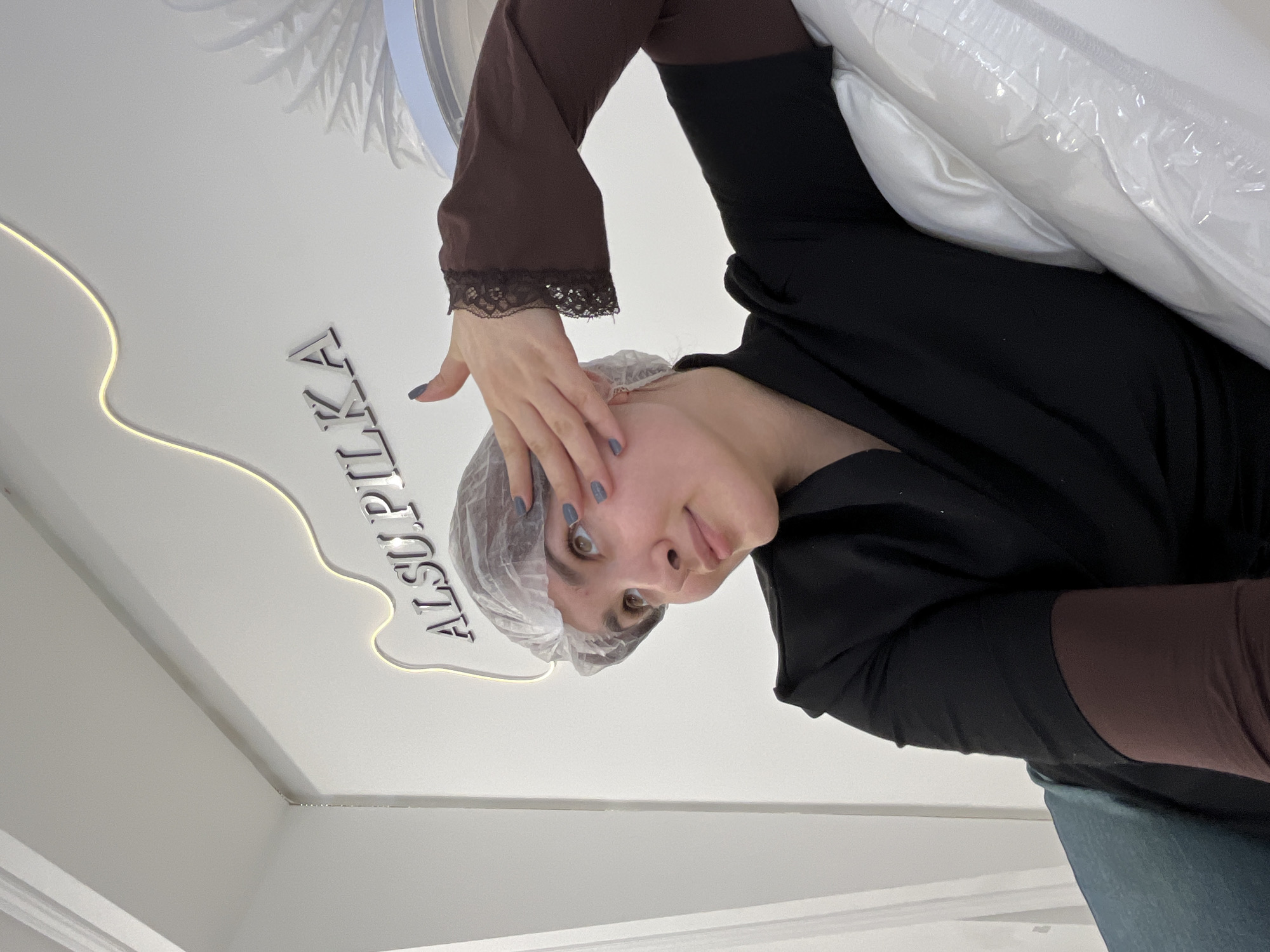 A young adult female is depicted in a beauty salon or aesthetic clinic identified as "ALSU.PILKA" at Lenina St, 32A, Neftekamsk, Republic of Bashkortostan, Russia. She wears a clear disposable hair cap, a dark brown long-sleeved undershirt with black lace cuffs, and a black short-sleeved top over it. Her fingernails are painted a muted blue-gray. She is seated or reclined, leaning against a white pillow or headrest wrapped in clear plastic. To her left, a dark teal upholstered surface is visible. Her right hand is raised with fingers splayed, touching or hovering near her right temple. She looks upward and to her right with a slight smile. The background consists of a plain white wall with silver dimensional letters spelling "ALSU.PILKA" and a wavy yellow LED light strip installed above the text. The scene is brightly and evenly lit.