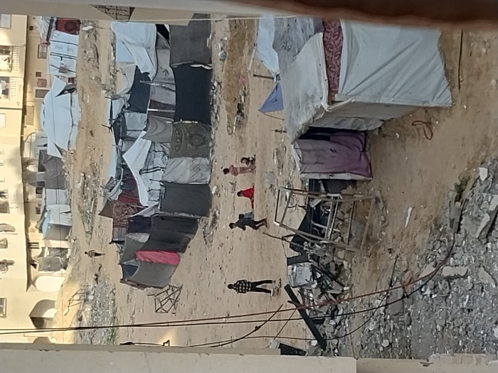 The image depicts a desolate, open sandy area in Khan Yunis, Palestinian Territory, serving as an improvised encampment amidst visible destruction. In the immediate foreground on the left, a substantial pile of grey concrete rubble, shattered building materials, and twisted rebar is present, with black electrical wires hanging loosely above it. A metal frame, possibly from a collapsed structure, stands upright within this debris.
The central and mid-ground consist of a large, flat expanse of light brown sand, sparsely dotted with smaller debris and mounds of dirt. Scattered across this area are numerous makeshift shelters, primarily constructed from tarpaulins, blankets, and fabric sheets of various colors (including white, dark grey, black, red, and patterned material) stretched over frames or poles. A prominent cluster of these shelters forms an irregular semicircle in the middle ground.
Approximately five children are visible in the central sandy area. On the left, one child wearing a plaid shirt and dark pants stands, while another child in dark clothing is captured mid-stride, moving towards the right. Further to the right, three younger children are near the ground: one in red clothing appears to be sitting or kneeling, another in pink clothing stands, and a third is partially obscured. In the far background, a single individual, likely a child, walks across a sandy elevation between the encampment and permanent structures.
Beyond the encampment, in the background, several multi-story concrete buildings of a residential nature are visible. Some exhibit arched ground-level openings. More makeshift shelters are dispersed closer to these permanent structures. The scene is illuminated by natural daylight.