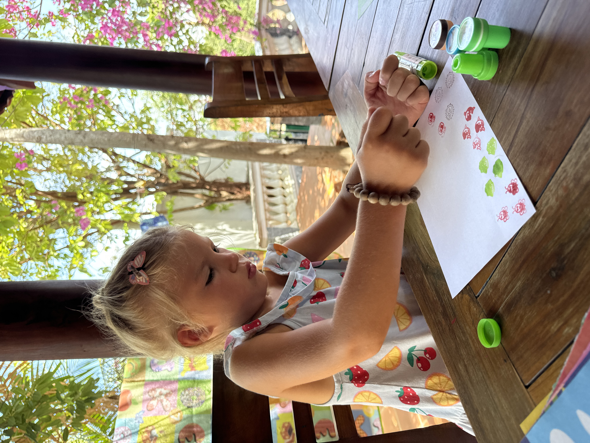 A young blonde girl sits at a wooden table using colorful stamp pads on a piece of paper. She wears a fruit-patterned dress and a bracelet. A butterfly clip adorns her hair. The table holds several stamp pads and a paper with stamped images of bugs and other motifs. The scene appears to be outdoors, judging from the foliage and sunlight visible in the background. Considering the location of Mũi Né, Vietnam, there may be tropical plants and trees in the background, perhaps a veranda or a patio setting.