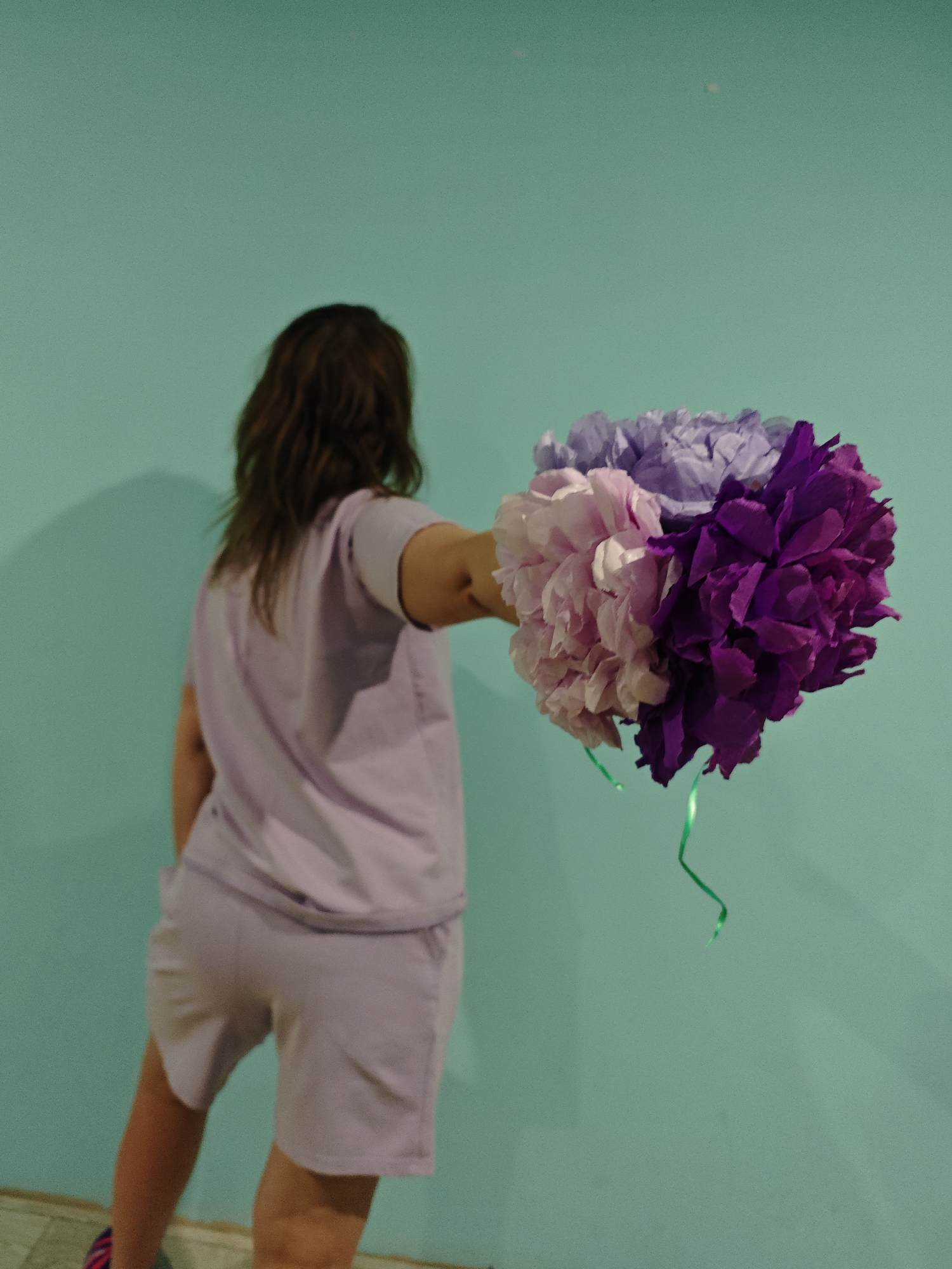 A person is standing indoors with their back facing the camera, holding a bouquet of paper flowers. The person has brown hair and is wearing a matching light purple short-sleeved shirt and shorts. The bouquet appears to be made of tissue paper in shades of pink, purple, and dark purple, with a green ribbon hanging down. The background is a solid aqua-colored wall. A portion of the floor is visible in the lower left corner. The location is not identifiable as Markovo, Russia based on the image's details.