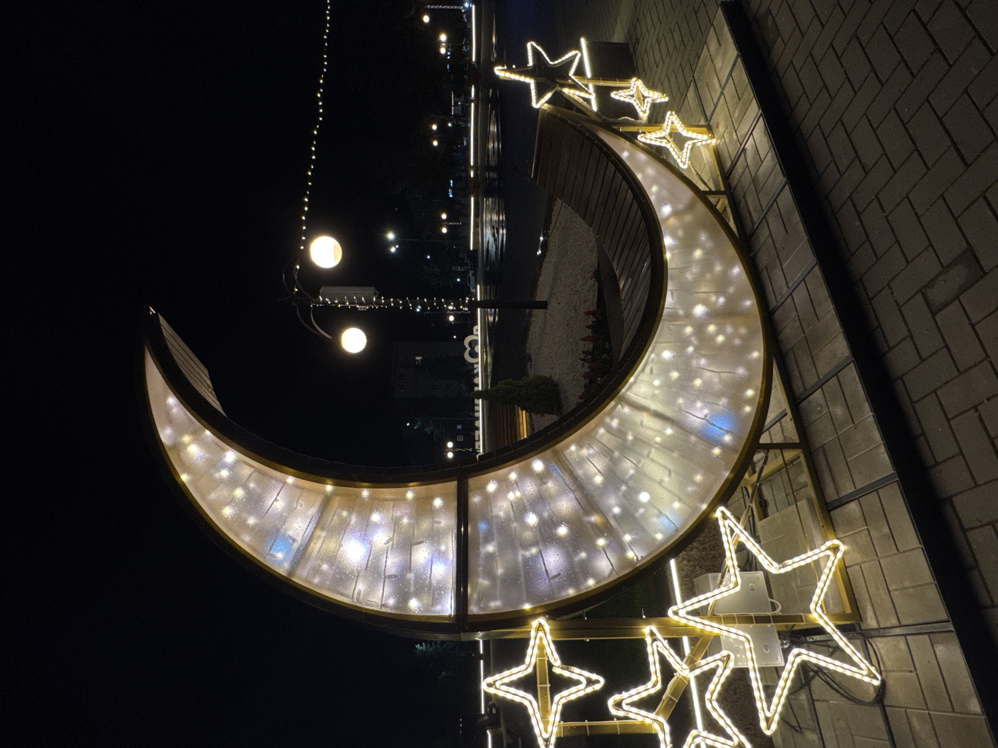 The image presents an illuminated outdoor scene at night in Karachayevsk, Russia. The primary subject is a large, crescent moon-shaped structure functioning as a decorative bench. This structure features a gold-colored metallic frame enclosing a translucent, frosted paneling that is internally lit by numerous white lights, creating a sparkling effect. A curved wooden bench is integrated into the concave side of the moon structure. Flanking the moon are several five-pointed star-shaped light decorations, outlined by white LED lights, standing on the paved ground. The ground is composed of light-toned rectangular pavers laid in a herringbone pattern. In the background, a lamppost with two spherical lights and wrapped string lights is visible. A horizontal string of small, warm white lights extends across the upper background. Further into the dark distance, additional streetlights illuminate distant areas, and some ground surfaces appear reflective. A small, conical evergreen shrub is situated behind the moon structure. No individuals are present in the scene.