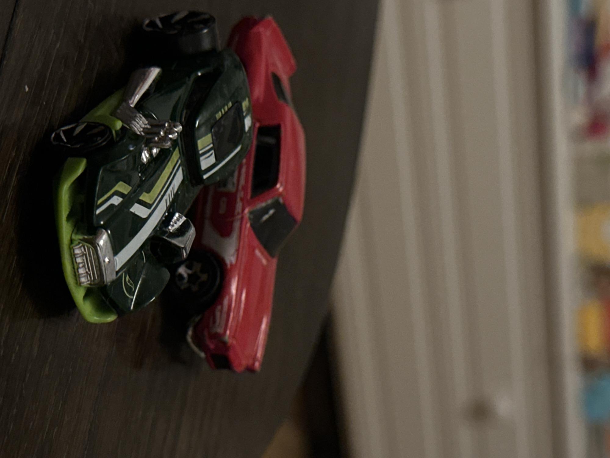 Two toy cars are on a wooden surface. The car in the foreground is dark green with lime green accents and white racing stripes. It has exposed silver engine parts and large black wheels with silver rims. The car in the background is red with white racing stripes and silver rims. The background is out of focus and appears to be a piece of furniture with various objects on top. There is no indication of the city of Sofia, Bulgaria.