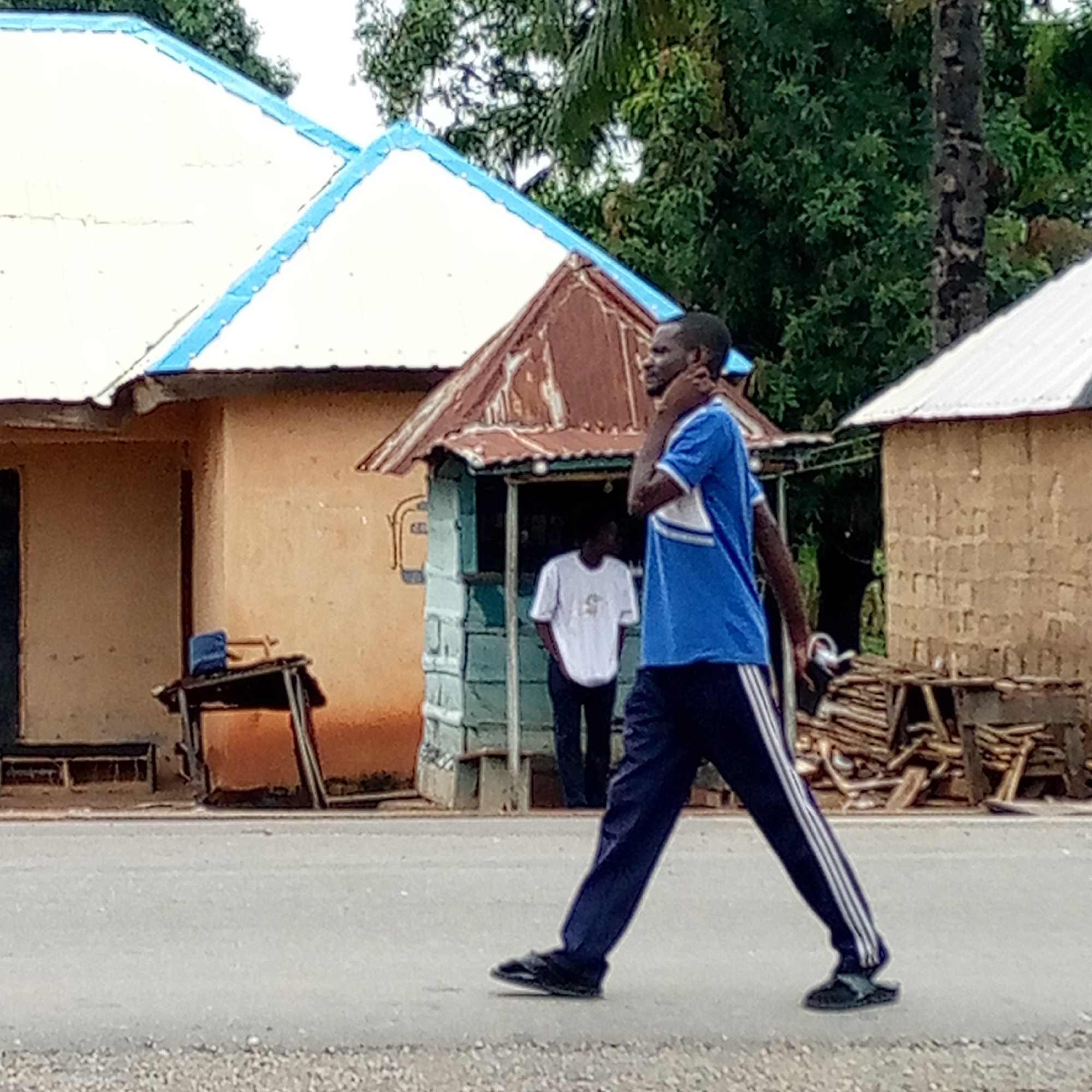 A man is walking across a paved road in Mararaba, Nigeria. He wears a blue t-shirt and dark-colored pants with white stripes. Another man is visible inside a small structure with a rusty roof and blue trim; this structure appears to be a small shop. Buildings with corrugated metal roofs line the road, and a stack of firewood or lumber sits near one building. Trees are visible in the background.