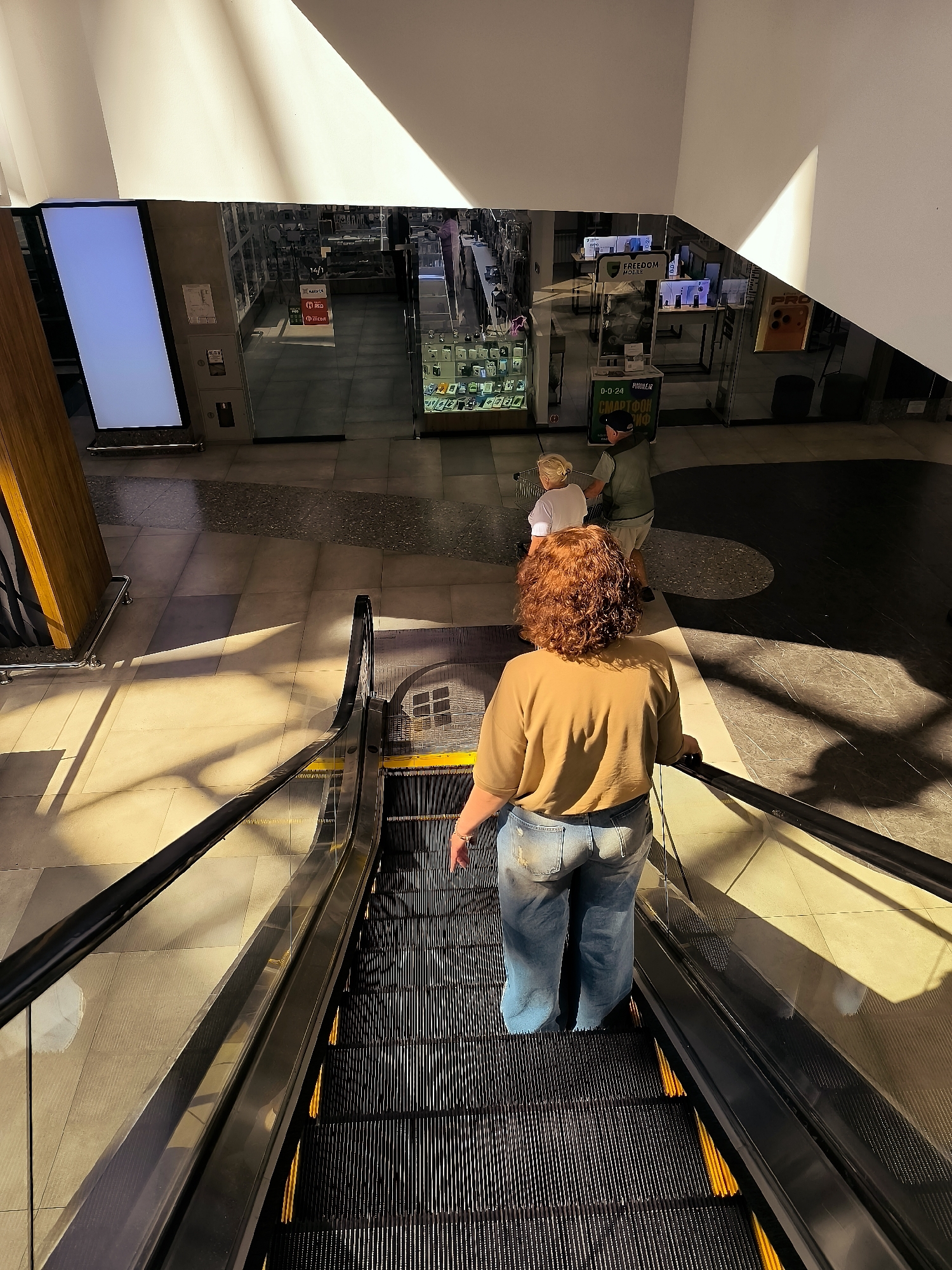 The image shows a woman with curly red hair, wearing a tan t-shirt and ripped light blue jeans, descending on a black escalator. She is positioned in the foreground, with her back to the camera. Her right hand rests on the escalator handrail.
Behind her, further down the escalator, is a young blonde child in a white t-shirt and a person wearing a green shirt and a dark baseball cap. They are also descending the escalator.
The escalator is located in an indoor space with tiled flooring. Harsh sunlight streams in from the upper left, casting distinct shadows across the floor and the escalator. In the background, through a large glass storefront, are displays of merchandise. The storefront has signage with text, including what appears to be "FREEDOM MOBILE". This scene is likely within a shopping mall or commercial building in Qarağandı city administration, Kazakhstan.