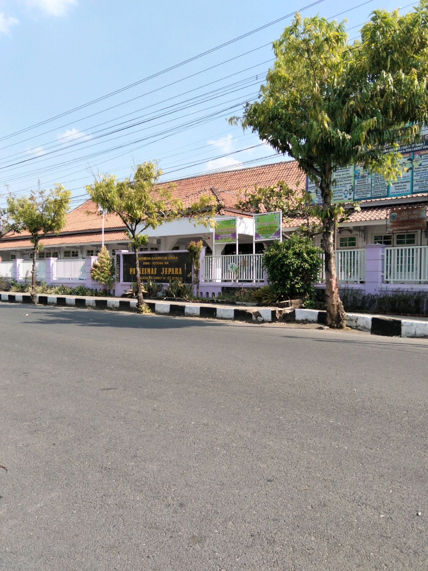 This is a street-level, medium-shot, eye-level photograph of the Puskesmas Jepara, a public health center in Jepara, Indonesia, located on a paved road under a clear blue sky with visible power lines overhead. In the foreground, a dark gray asphalt road occupies the bottom half of the frame, with a white and black striped curb on the right side. Behind the curb is a sidewalk and a low, white fence topped with a purple accent. Several trees with green foliage are planted along the sidewalk.
The Puskesmas Jepara building is visible in the midground, a single-story structure with an orange-tiled roof and white walls. A prominent sign in front of the building reads "PEMERINTAH KABUPATEN JEPARA DINA - KESEHATAN DAN PUSKESMAS JEPARA." Multiple smaller signs and posters are affixed to the building's facade and fence. To the left of the main entrance, there is a sign with a green cross and text that says "KOPPI REHAT." Another large tree stands in front of the building, partially obscuring it. The overall scene is well-lit by sunlight, casting shadows and highlighting the vibrant green of the trees and the blue of the sky. There are no people visible in the image.