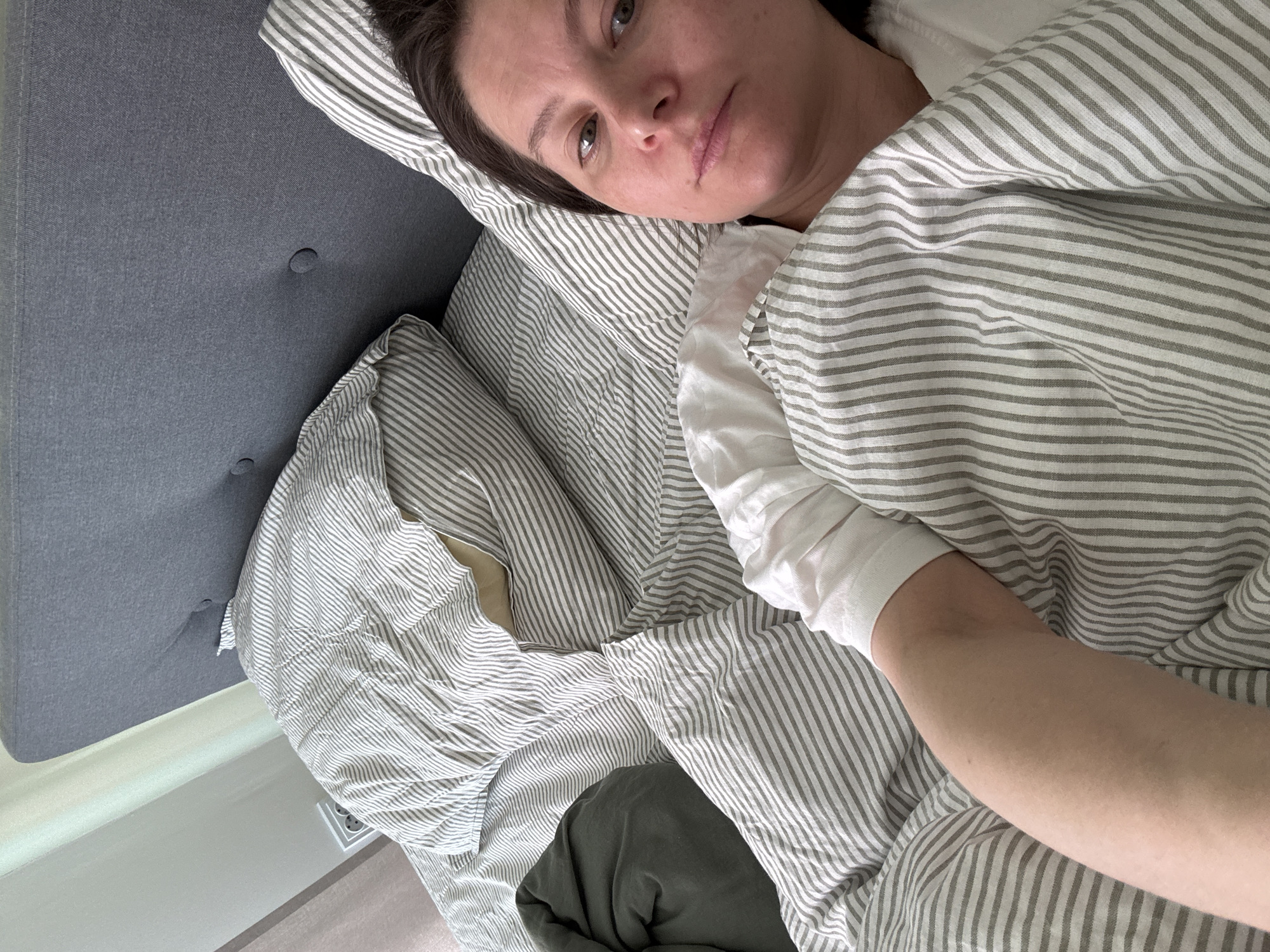A woman lies in a bed with striped bedding. Her head rests on a pillow with similar striped fabric. She is partially covered by the bedding and appears to be wearing a white top. Her arm lies extended on top of the bedding. A gray headboard is visible on the left side of the image. The location in the city of Люк, Russia is not discernible from the image.