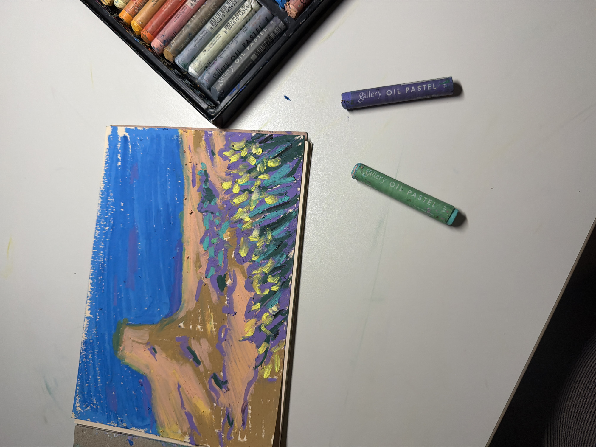 The image depicts an overhead view of a white table displaying a partially completed oil pastel drawing of a landscape, along with a box of oil pastels and two individual oil pastel sticks. The drawing, rendered on a light brown paper pad, features a prominent mesa-like formation in the background under a bright blue sky with hints of purple clouds. The foreground is a vibrant mix of green, yellow, and purple strokes representing vegetation. To the right of the drawing is a black case filled with a variety of oil pastels, showcasing rows of colorful sticks. Two individual oil pastel sticks, one green and one purple, are placed on the table in front of the drawing. The scene is lit, casting subtle shadows on the table. The image does not contain any identifiable elements that would suggest a specific location within Dongducheon-si, South Korea, or any other city.