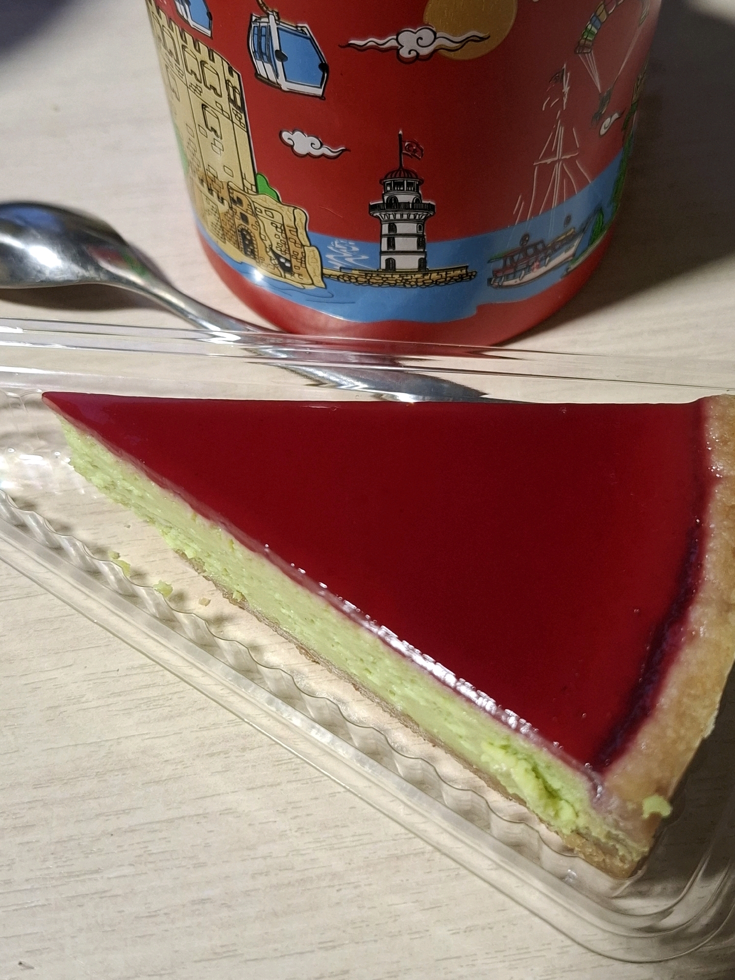 A slice of cheesecake with a red jelly topping sits in a clear plastic container on a light-colored, wood-grain surface. To the left of the container, a silver spoon rests diagonally. Behind the cheesecake and container, a red mug with various illustrations, including a lighthouse, a boat, and a cable car, is visible. The image is taken from a high angle, focusing on the dessert. There is no indication of the city of Tyumen, Russia, in this image.