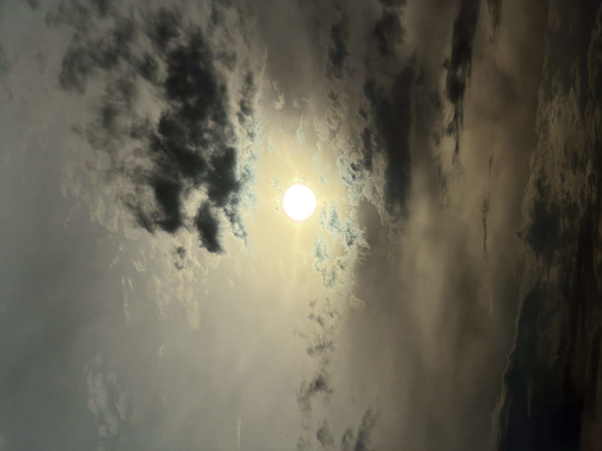 The image captures a dramatic sky over Mramor, Bulgaria, dominated by the sun peeking through a dense, variegated cloud cover. The sun, a bright, luminous disc, is positioned slightly off-center to the left, casting a strong yellow-white glow. Dark, heavy clouds, tinged with grey and brown, swirl around the sun, creating a sense of depth and atmospheric intensity. Some lighter, wispy clouds are also present, interspersed with the darker formations. The overall color palette is muted, with shades of grey, brown, and yellow, suggesting either dawn, dusk, or an overcast day with intermittent sun. There are no discernible people, structures, or other objects in the frame; the scene is solely focused on the celestial elements. The image appears to be a telephoto shot, emphasizing the textures and formations of the clouds and the brilliance of the sun.