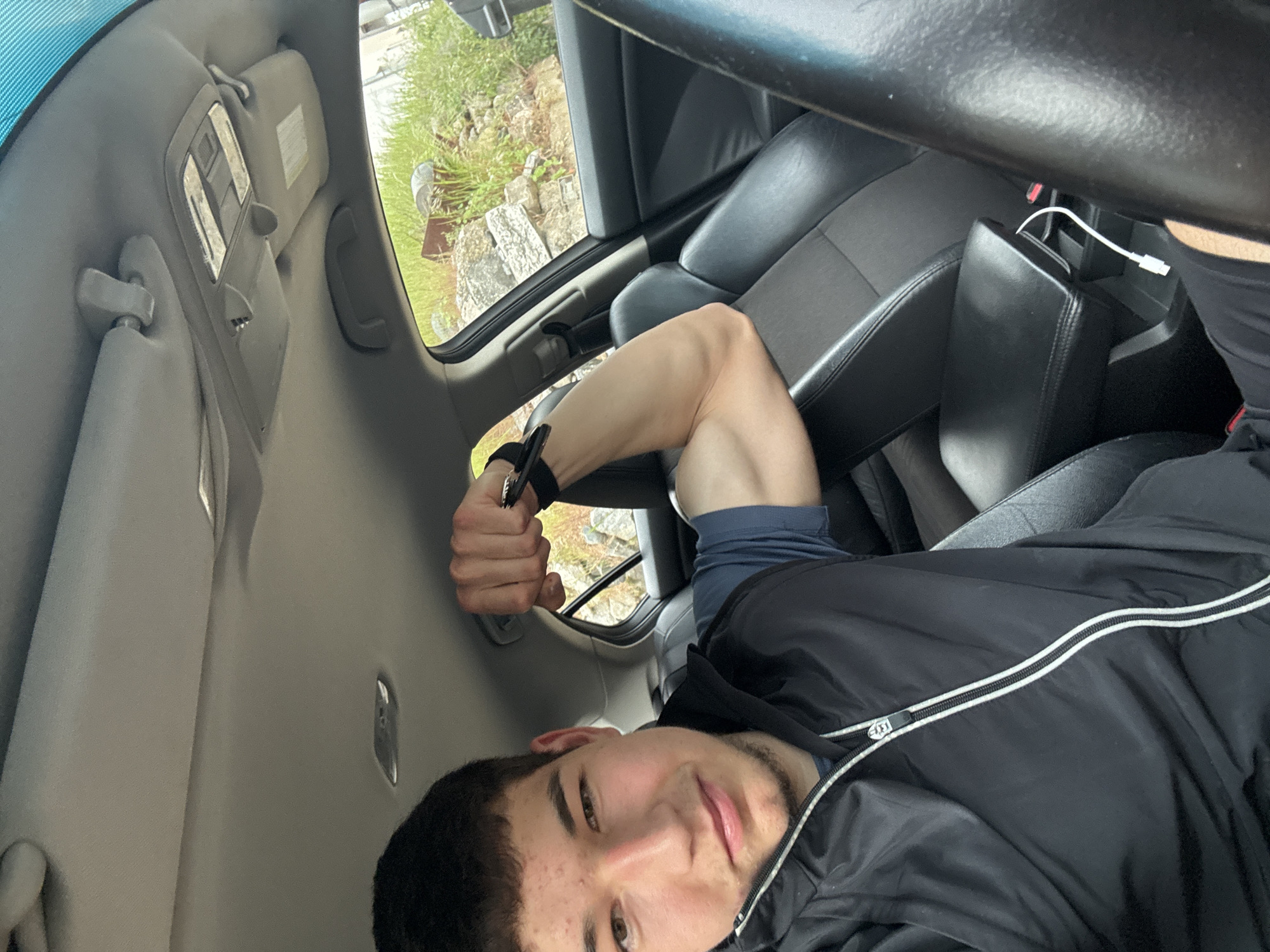 A young man is sitting in the driver's seat of a car, posing for a selfie. The car's interior includes gray seats, a black dashboard, a steering wheel, and a center console with a charging cable plugged in. Outside the car window, there are green plants and stone structures. The photograph appears to be taken during daylight hours. The image location is not in Zlatograd, Bulgaria.