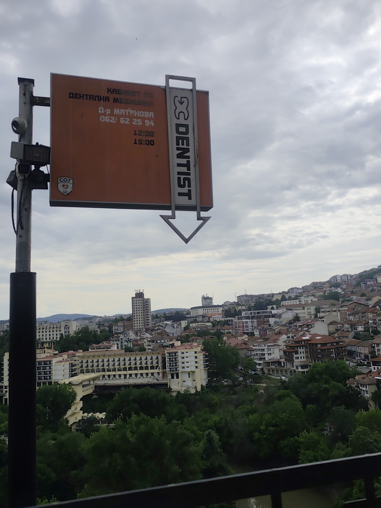 The image shows a view of a city with a cloudy sky in the background. In the foreground, there's a sign on a pole for a "DENTIST" (written vertically) with the information "КАБИНЕТ По ДЕНТАЛНА МЕДИЦИНА" (Cabinet of Dental Medicine), "д-р МАТЪШОВА" (Dr. Matshova), the phone number "062/62 25 94", and the hours "12:00-19:00". The location appears to be in the city of Veliko Tarnovo, Bulgaria. The city is spread across hills, with various buildings, trees, and a river running through the scene.