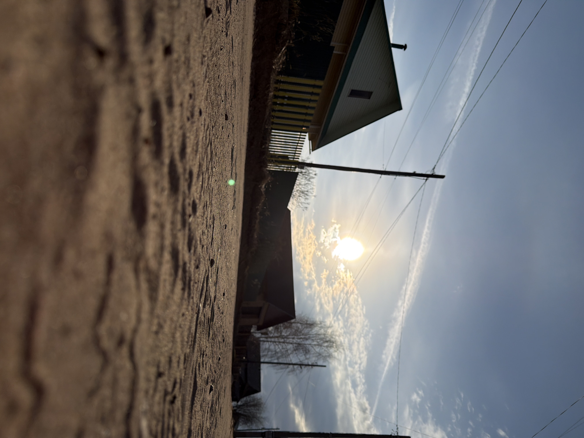 The image captures a rural or semi-rural landscape from a low-angle perspective, looking up towards the sky. The foreground is dominated by a textured ground surface, likely dirt or gravel, showing various shades of brown and small, dark specks.
In the midground, several buildings are visible. One prominent structure on the right side features a light-colored facade, possibly striped in white and green, with a dark, peaked roof and a small, rectangular window. Behind it, other buildings and bare trees or shrubs can be seen. A dark utility pole with several power lines stretches across the scene, extending upwards towards the sky.
The background is a partly cloudy sky with a bright, intense sunburst visible through a patch of white, scattered clouds in the upper-middle portion of the frame. The sunlight casts a warm glow, suggesting either early morning or late afternoon. Long, thin contrails or wispy clouds are also present across the blue sky.
No people or visible text are present in the image. The scene depicts a tranquil outdoor setting in Chaikovsky, Russia.
