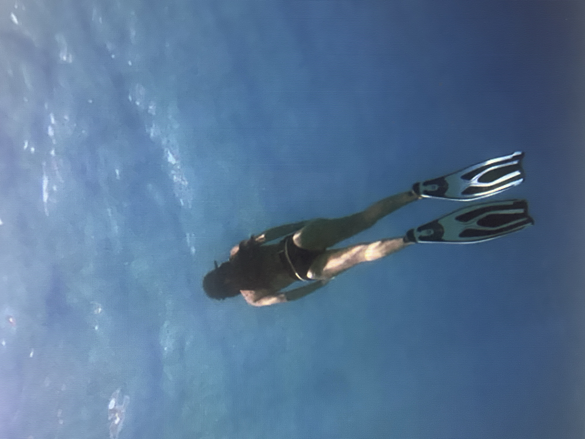 This media file depicts a person free-diving underwater. The subject, wearing dark swim trunks and black-and-white swim fins, is positioned horizontally in the frame, facing towards the left. Their body is streamlined, suggesting they are in motion through the water. The fins are prominent, with a distinctive black and white pattern.
The setting is an underwater environment, characterized by clear blue water. The lighting suggests a daytime scene, with light filtering down from the surface, creating subtle variations in the water's color and texture. Some lighter, cloud-like patterns are visible in the water, possibly indicating the presence of sediment, plankton, or the play of light and shadow.
The activity is clearly free-diving, a form of underwater swimming using breath-hold techniques. The subject's posture and the equipment (fins) indicate an intentional movement through the water, likely for recreation or exploration. There are no visible text elements in the image. The overall impression is one of calm and immersion in the aquatic environment.