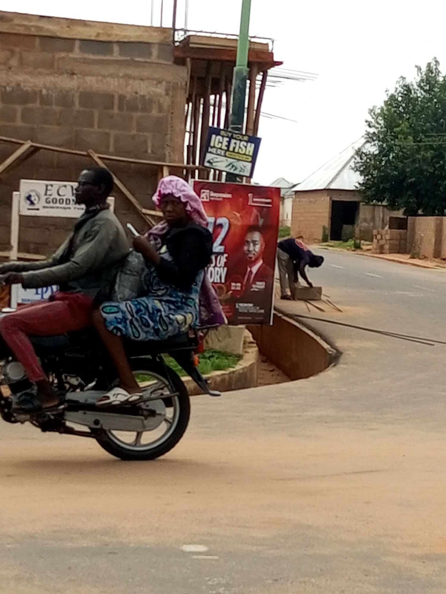 Two individuals are riding a motorcycle on a paved road in Tempure, Nigeria. The driver, a man wearing glasses and a dark jacket, is looking forward. The passenger, a woman wearing a pink head covering and a blue and white patterned dress, is holding a mobile phone and looking to the side. In the background, a construction site is visible with brick walls and scaffolding. A large poster with an image of a man in a suit and text reading "2 SOF ORY" is also present. Another smaller sign displays "BUY YOUR ICE FISH". A third person is seen bending down near the road, appearing to interact with some objects. Trees and buildings line the street. The weather appears overcast.