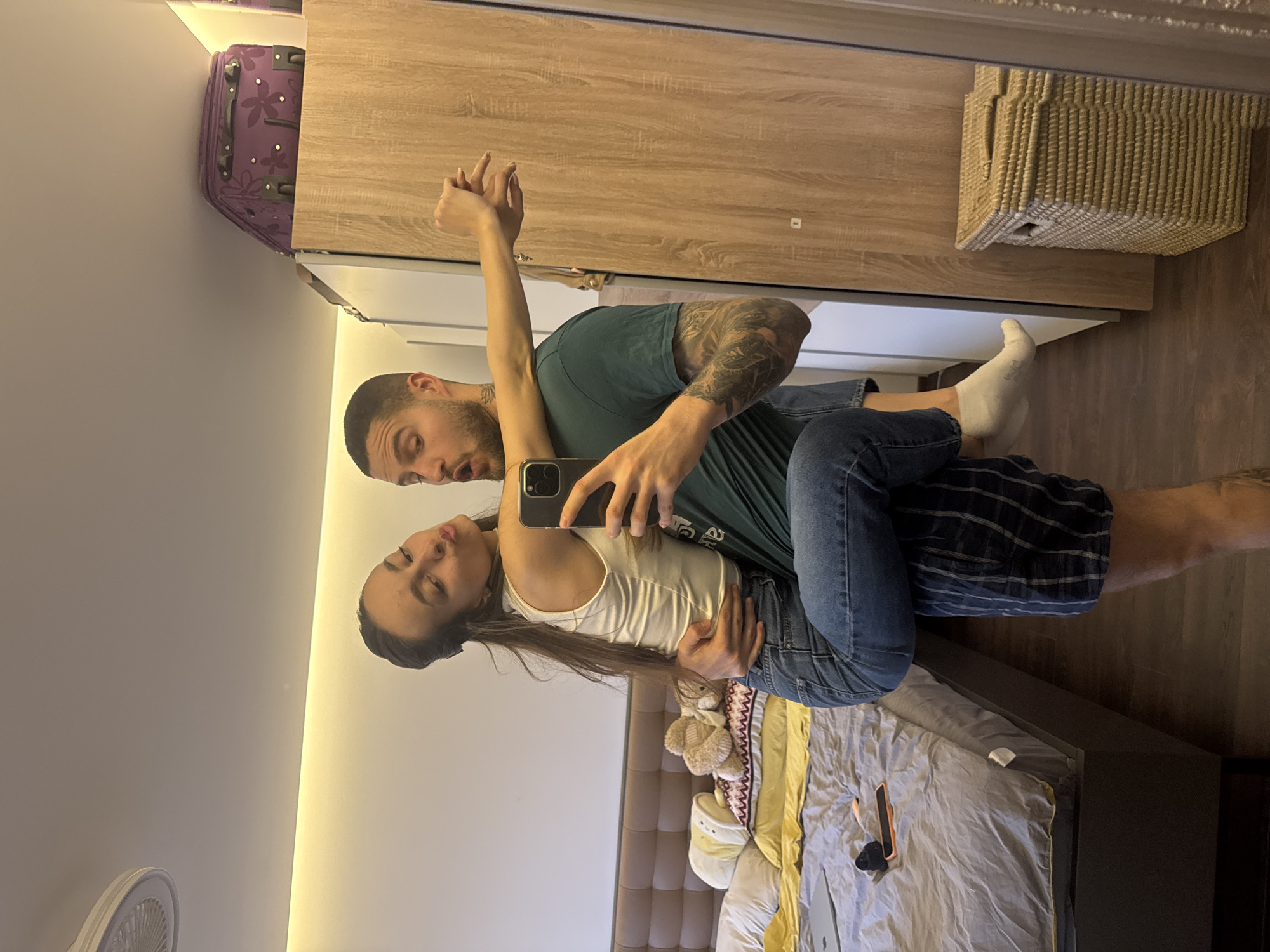 A mirrored image shows a man and a woman embracing and taking a selfie in what appears to be a bedroom. The man is wearing a green t-shirt and blue jeans, and has tattoos on his arm. The woman is wearing a white top and blue jeans. They are both looking at the phone. In the background, there is a bed with a grey and yellow duvet, a wardrobe, a suitcase, and a woven basket on top of the wardrobe. The location appears to be an interior setting, potentially a room in a residential building, the city of Sofia, Bulgaria. A fan is visible on the wall.