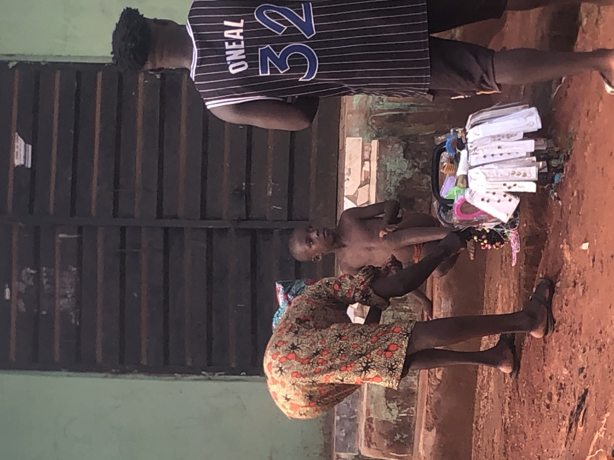 This image captures an outdoor market scene in Ukwu-Abwa, Nigeria, under daylight conditions. The ground is unpaved, a reddish-brown dirt surface.
In the foreground, a person with dark skin is seen bending over, facing away from the viewer. This individual is wearing a colorful, patterned garment, possibly a dress or skirt, featuring orange, green, and black designs. They have dark legs and are wearing black flip-flops.
To the right, a young, shirtless child with dark skin is crouching or sitting on the ground, looking towards the camera. Behind them, a taller individual with dark skin stands upright, wearing a dark-colored, vertically striped jersey. The back of the jersey clearly displays the name "O'NEAL" and the number "32" in blue text. This person has dark, textured hair, possibly dreadlocks.
Between the bending person and the standing individual, a small vendor's display is set up on the ground. It features a collection of various small items, predominantly hair accessories like combs, hair ties, and clips, many of which are individually packaged in clear plastic sleeves and arranged in a container.
The background consists of a dark, slatted structure, possibly a fence, shutter, or part of a building, extending vertically across the scene. The overall setting suggests a casual, local marketplace where goods are displayed for sale.