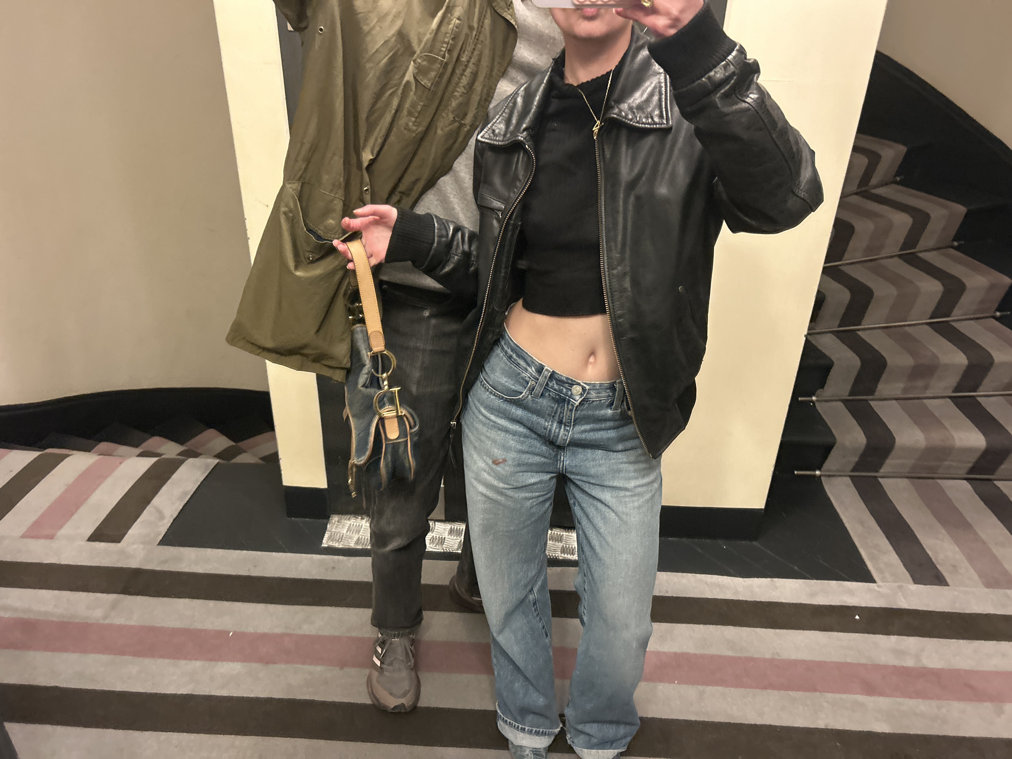 A person is posing for a photo in what appears to be a hallway or landing near a staircase. The person is wearing a black cropped top, blue jeans, and a black leather jacket. They are also holding a small handbag with a tan strap. A second person, partially visible, is standing to the left of the first person, wearing gray pants and shoes and standing next to a green coat. The staircase in the background has striped carpeting in shades of black, gray, and pink. The walls are painted in a light color. The location is Unknown, Unknown.