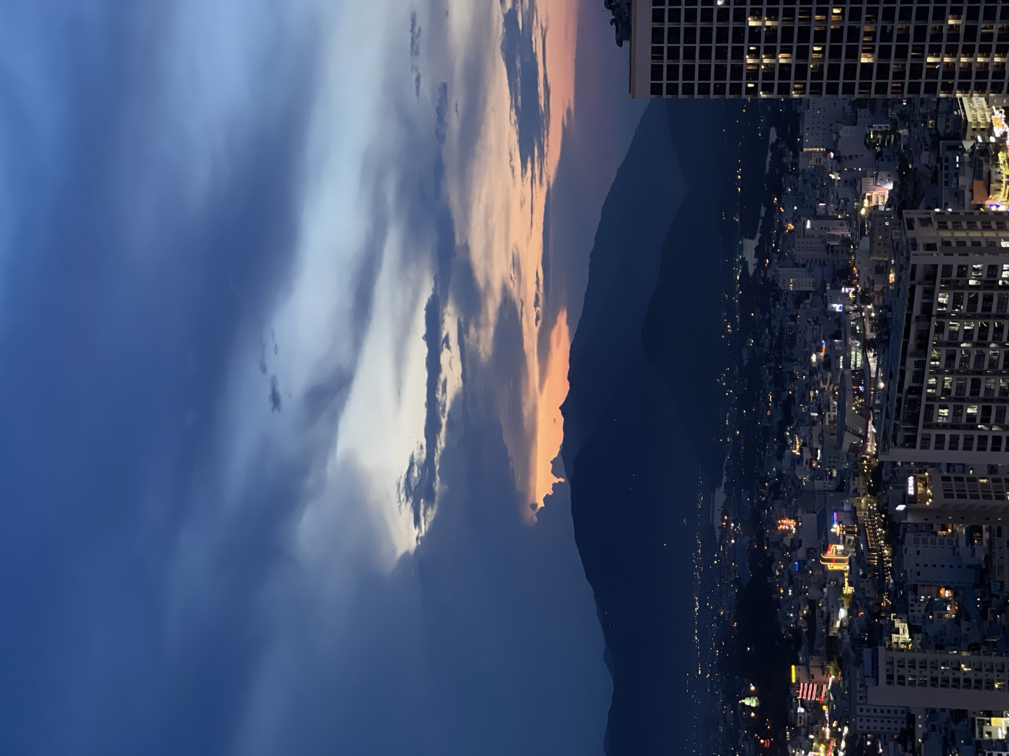 The image shows a nighttime view from a high vantage point in Nha Trang Ward, Vietnam. The foreground is dominated by a darkened mountain range, with a city skyline in the background illuminated with artificial lights from buildings, roads, and other infrastructure. A tall building stands close to the observation point. Above the mountains are clouds, some illuminated in orange and pink hues, and a generally blue sky. There are no people visible.