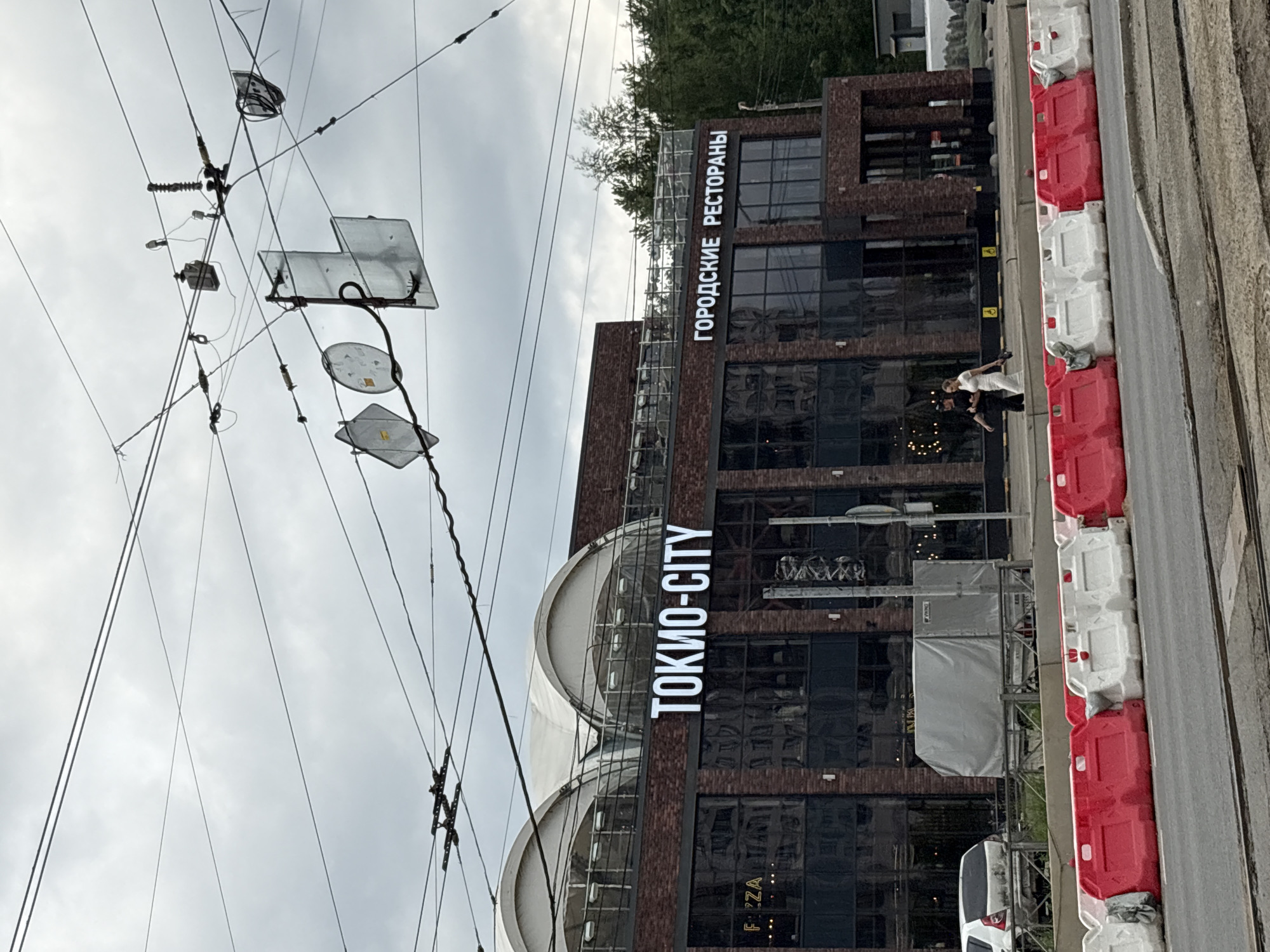 This is a medium shot, eye-level, outdoor photograph taken in the city of Imeni Morozova, Russia. The image captures a building with large illuminated signs that read "ТОКИО-CITY" and "ГОРОДСКИЕ РЕСТОРАНЫ." The building is constructed with dark brick and features large, dark-paned windows, and a modern, arched white roof structure. In front of the building, there are red and white plastic barriers lining a street. A white car and a gray covered trailer are parked behind the barriers. In the foreground, there are tram tracks. Two people, a man and a woman, are walking away from the camera on the sidewalk in front of the building. The sky is overcast and cloudy.