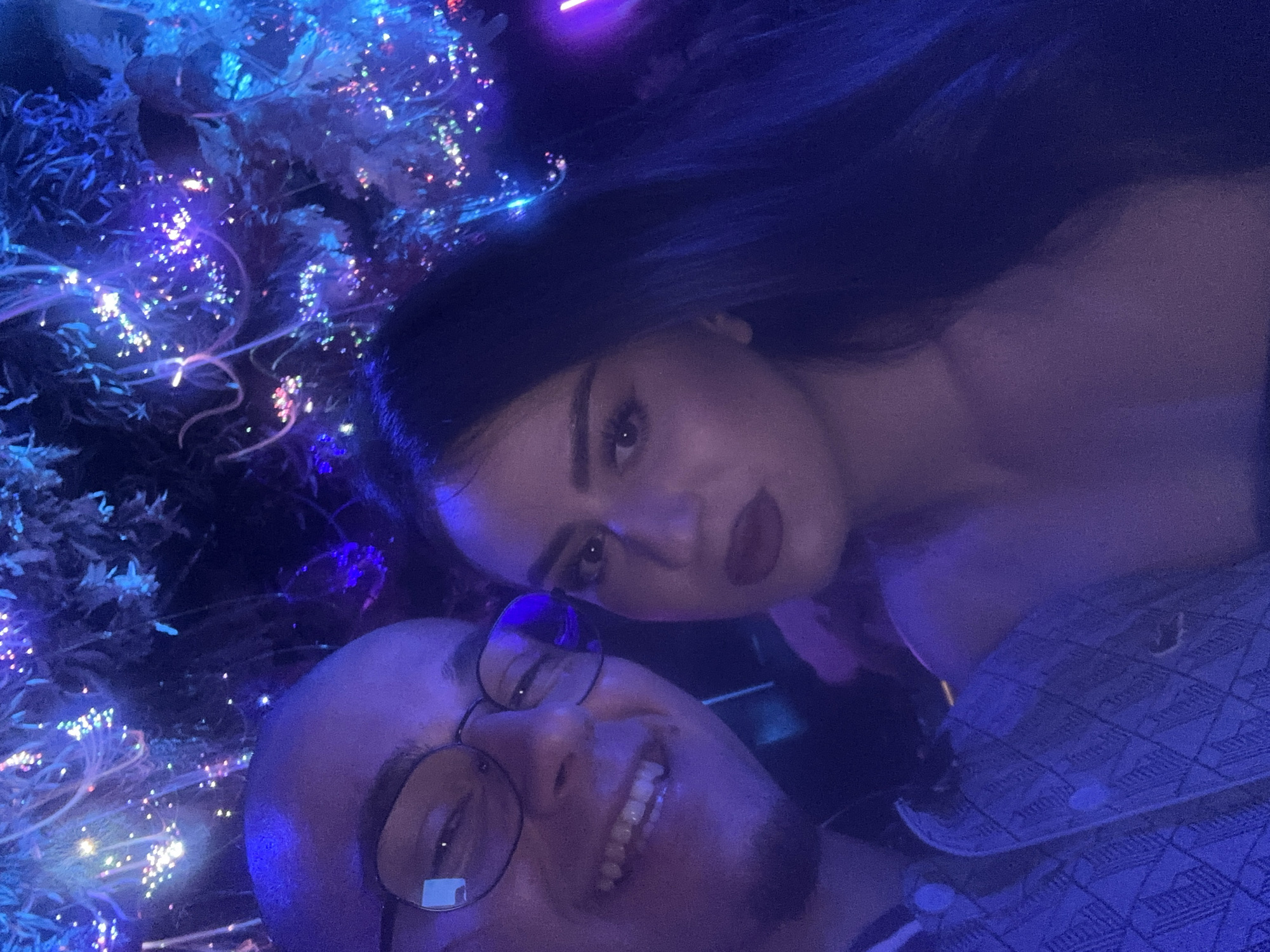 This vertical selfie features two individuals, a man and a woman, positioned closely together in a vibrant, artificially lit setting. The man occupies the lower foreground, with his head tilted upwards and to the right. He has a shaved head, wears clear-rimmed glasses, and is smiling broadly, revealing his teeth. His blue patterned shirt, possibly quilted or textured, is visible in the bottom right corner.
Behind and slightly above him is a woman, looking directly at the camera with a more neutral expression and dark red lipstick. She has long, dark hair and appears to be wearing a dark top. Her face is illuminated by the ambient blue and purple light.
The background is dominated by an array of bright, sparkling lights, predominantly blue and purple, with smaller hints of pink, green, and white. These lights appear to be embedded in or intertwined with dark foliage or decorative elements, resembling fiber optic displays or intricate LED installations, creating a fantastical, almost otherworldly atmosphere. The overall lighting scheme is dark, suggesting a nighttime setting, possibly within a club, bar, or an illuminated art exhibit in Sofia, Bulgaria. There is no visible text in the image. The mood conveyed is one of casual enjoyment in a lively, visually stimulating environment.