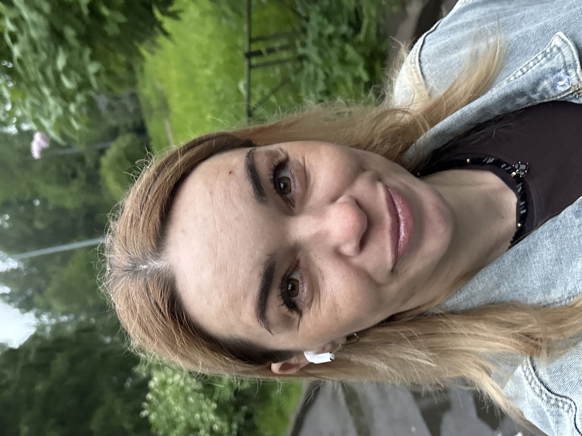 A woman with blonde hair and brown eyes is taking a selfie outdoors. She is wearing a denim jacket and a dark shirt with a beaded necklace. She has white earbuds in her ears. The background is a blurred view of trees and foliage, suggesting a park or natural area. The ground appears wet, possibly from rain.