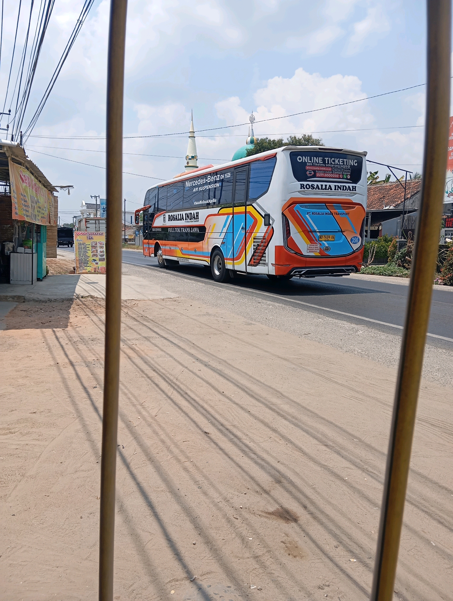 In Leman Benawi, Indonesia, a double-decker bus with the livery "ROSALIA INDAH" is seen driving on a paved road. The bus, a Mercedes-Benz OH 1626 with air suspension, displays "ONLINE TICKETING" on its rear. It is one of several vehicles visible in the urban environment, with houses and a mosque with a green dome also present. The scene is captured from behind vertical bars, suggesting a confined viewing perspective, possibly from a window or a barred structure. The ground in the foreground is dirt and gravel, with shadows cast by power lines and the bars. The sky is partly cloudy with a bright, daytime atmosphere. A food stall or small shop is visible on the left side of the image.