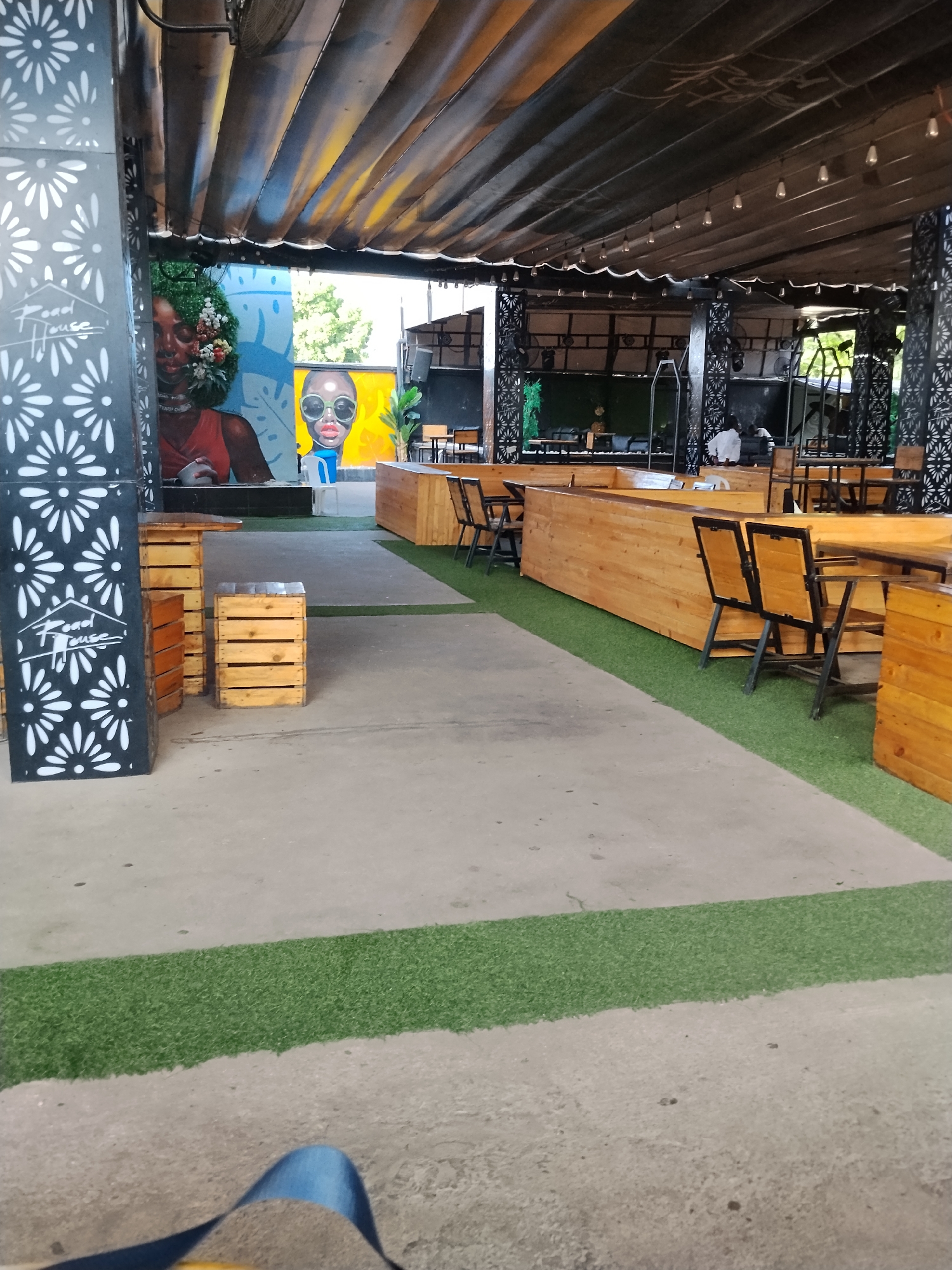 The image depicts the outdoor seating area of a restaurant or bar. The setting appears to be in Howayi, Nigeria, as suggested by the presence of what looks like a mural of a woman with a floral headdress and a depiction of a person wearing sunglasses. The area is covered by a fabric awning supported by pillars. Several wooden tables and chairs are arranged on a concrete floor, with a strip of artificial grass bordering the seating area. In the background, more seating and structures are visible, along with some people, including one person wearing a white garment, possibly a server. The overall ambiance is casual and open-air.