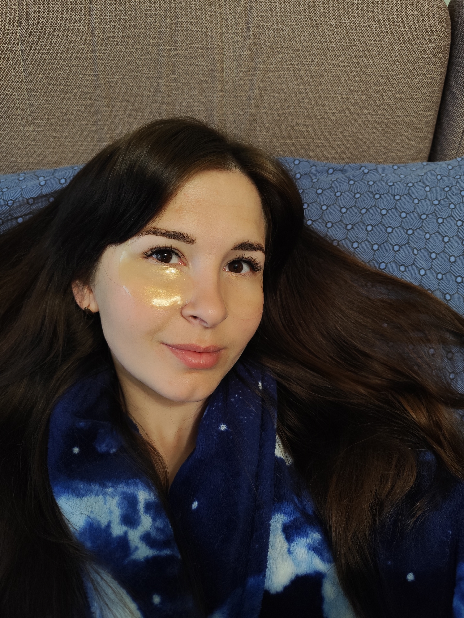 A woman with long, dark brown hair lies on a blue patterned pillow, wearing a gold eye patch under her left eye and a dark blue robe with a white tie-dye pattern. The background includes a beige couch. The location is not discernible as Фоки, Russia.