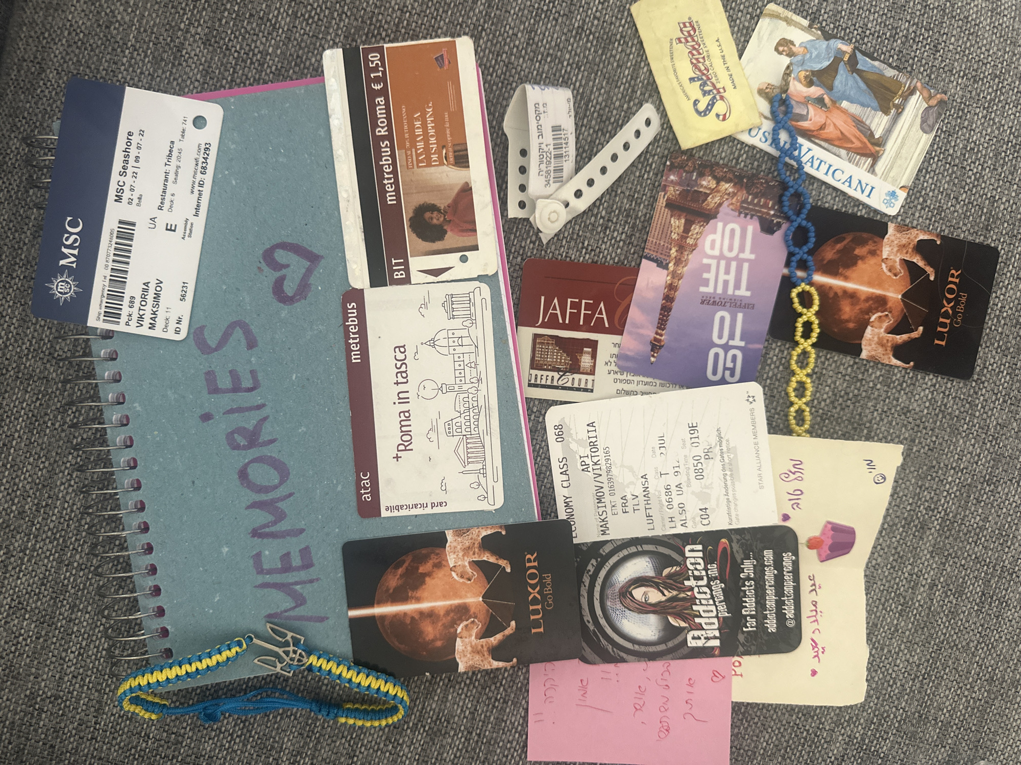The image shows a collection of travel memorabilia scattered on a gray surface. Dominating the scene is a light blue spiral notebook with "MEMORIES" written on it in purple. Overlapping the notebook is an MSC cruise ticket, a "metrebus Roma" ticket, and a blue and yellow Ukrainian bracelet. Other items include metro tickets from Rome, Italy; a "Jaffa" card; "Luxor" cards; a "Go to the Top" Eiffel Tower card; a card depicting Vatican art, a Splenda packet; a receipt; a "Addiction Piercings" card; and a small card with Arabic writing. The photograph's composition and content suggest the collection is personal and likely related to travel experiences of Viktoriia Maksimov. There is no information in this image to determine a location in the city of Petah Tikva, Israel.