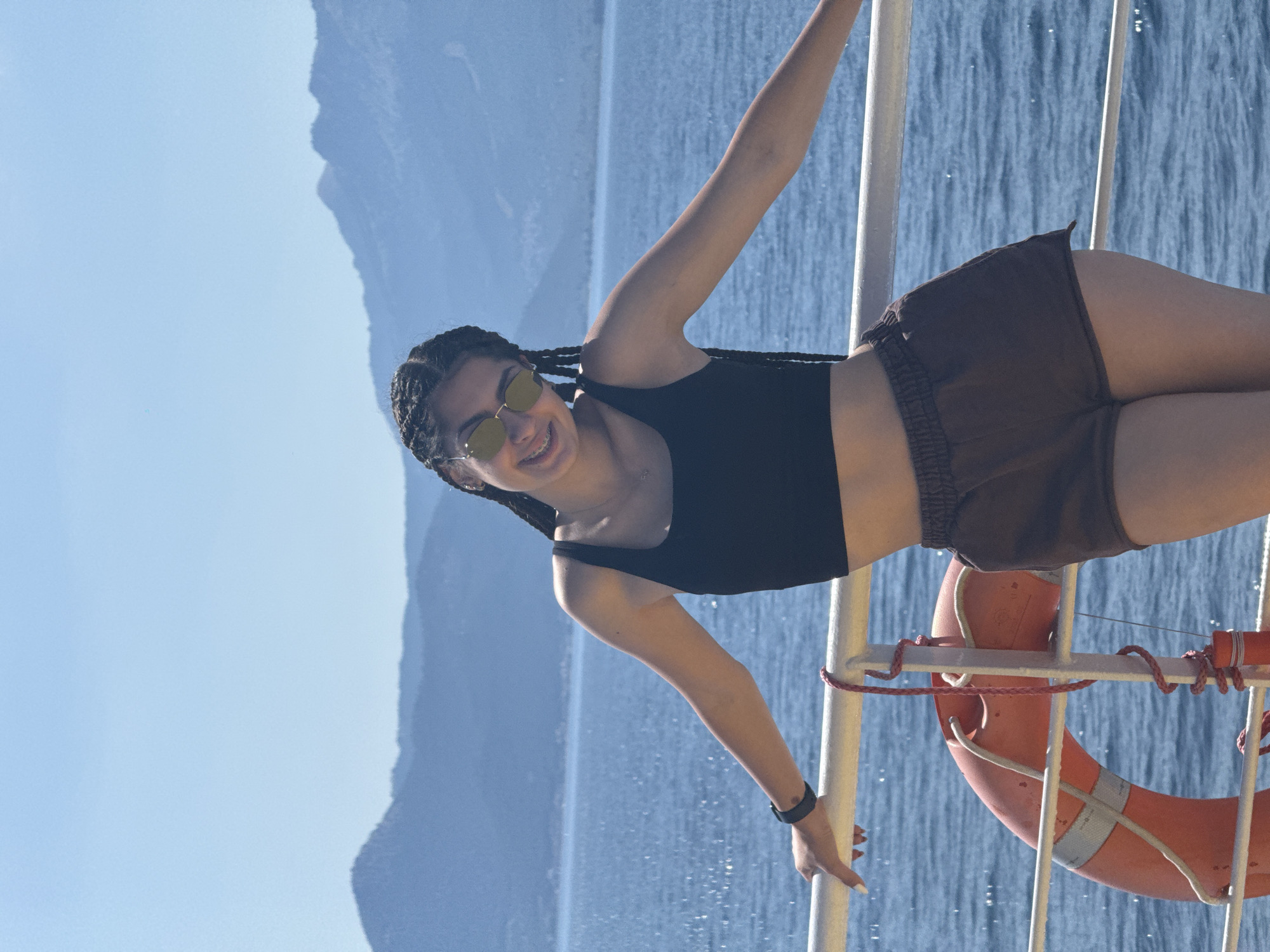 A young woman with braided hair and sunglasses stands on the deck of a boat, leaning on a railing. She is wearing a black crop top and brown shorts. Behind her, the sea stretches out to a distant, mountainous coastline under a clear blue sky. A life preserver is visible on the deck. The location is identified as an unspecified place in Greece.
