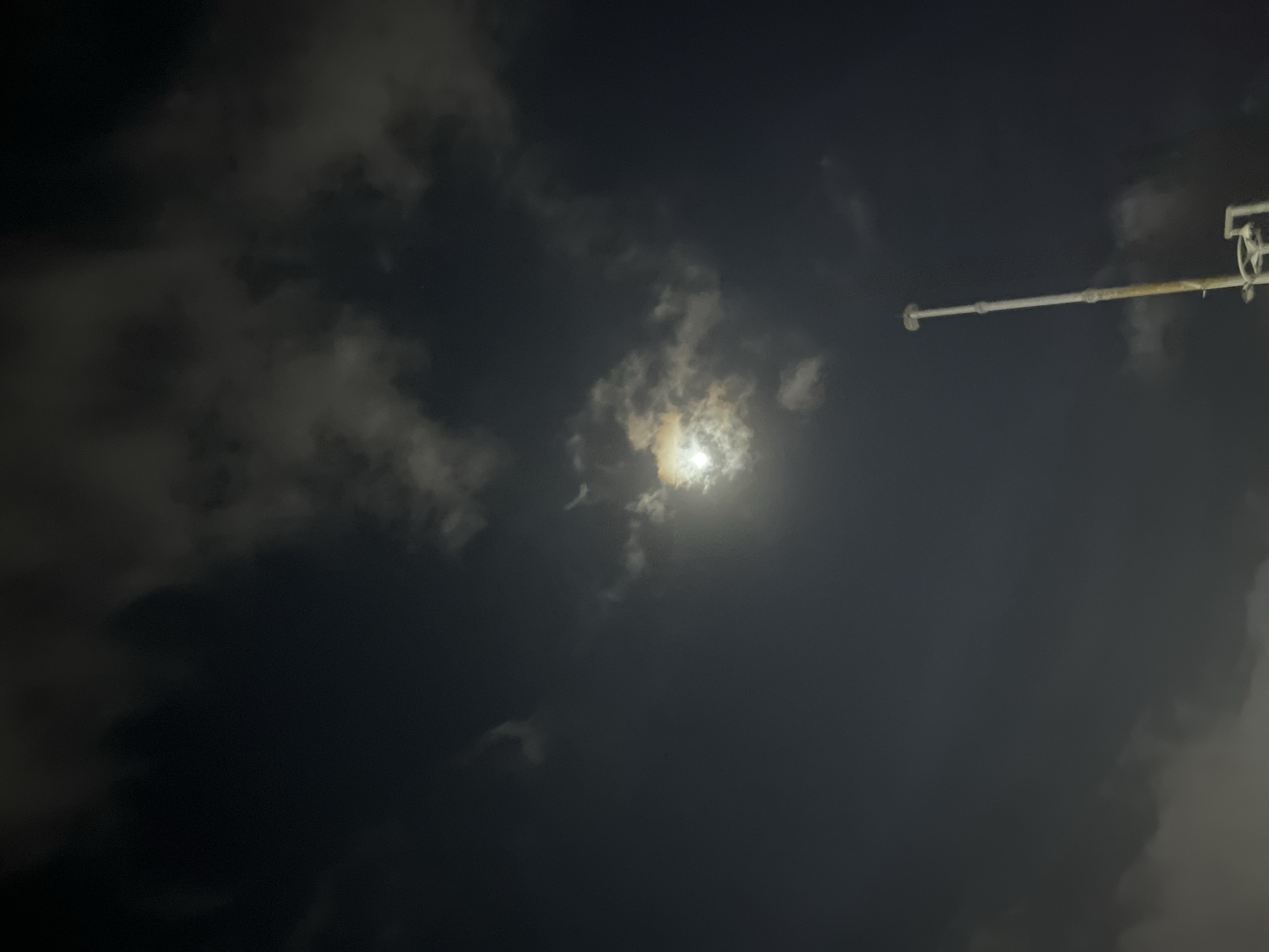 The image depicts a nighttime scene in Denpasar, Indonesia, dominated by a dark, cloudy sky. A bright, partially obscured moon shines through the clouds, casting a soft light. In the foreground, a tall, white pole with some rust near its base extends upwards, suggesting it might be part of a lighting fixture or antenna. The lower portion of the pole reveals some mounting hardware. The overall atmosphere is dimly lit and somewhat eerie, characteristic of a cloudy night.