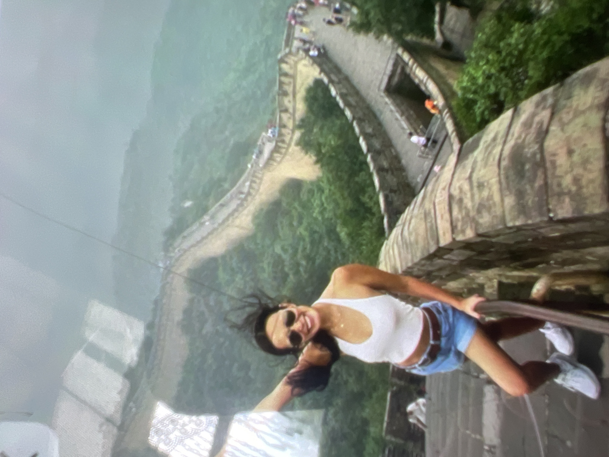 The image depicts a woman standing on stone steps, holding a white cloth in her raised left hand. She is wearing a white halter-neck top, denim shorts, and white sneakers. The woman has dark hair and is wearing sunglasses and a necklace. She is smiling and looking towards the camera. Behind her, the Great Wall of China stretches across a misty, mountainous landscape covered in lush green trees. Other tourists are visible walking along the wall in the distance. The environment appears to be a historical landmark. The image does not show the city of Kara-Dıykan, Kyrgyzstan.