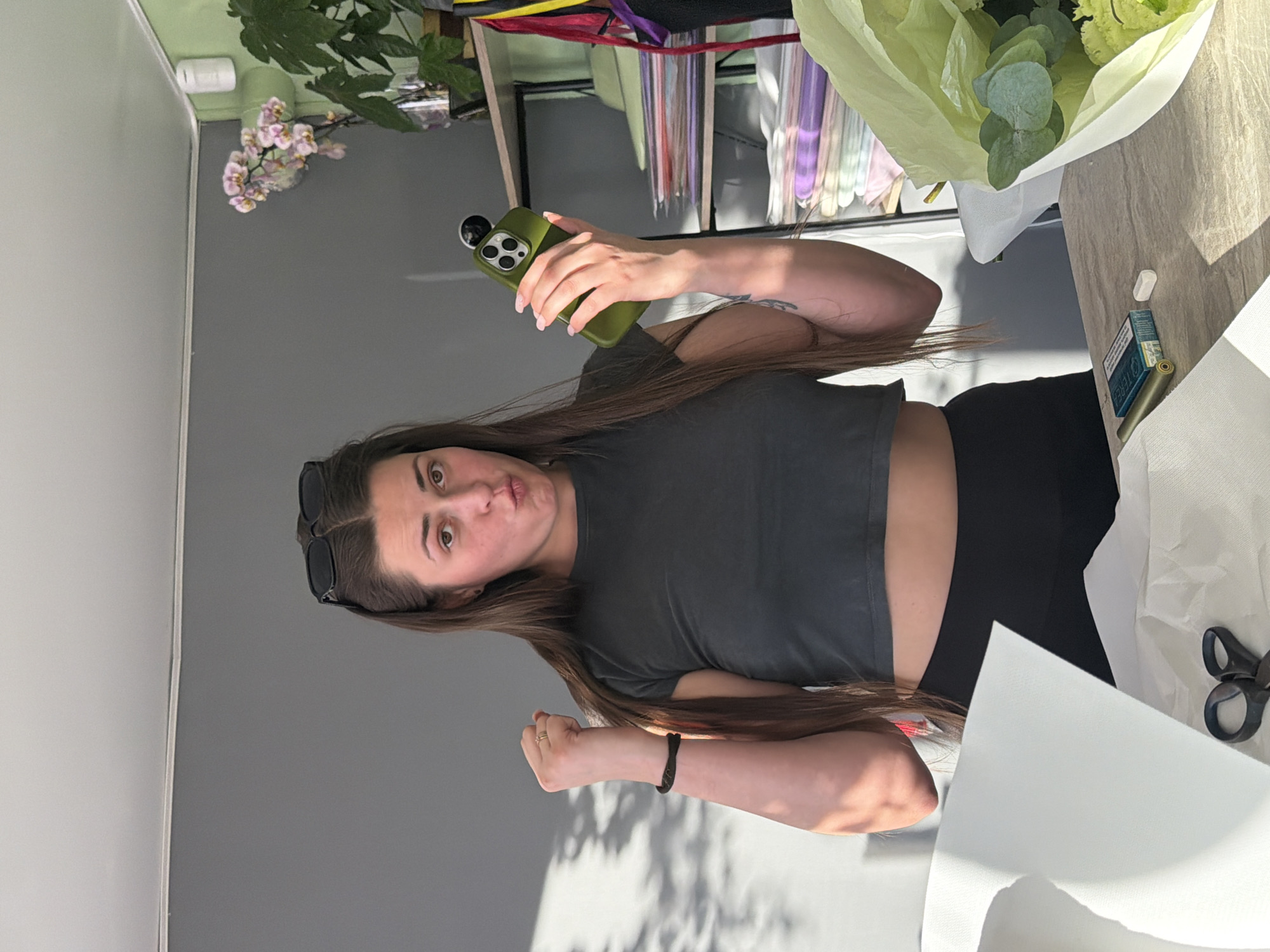 A medium shot shows a woman with long brown hair, wearing a dark grey crop top and black leggings, holding a green iPhone in her right hand. She stands in an indoor setting, possibly a workshop or studio, with a grey wall behind her. Sunlight casts shadows on the wall. In the foreground, a wooden table holds a bouquet wrapped in light green paper, white paper, a pair of black scissors, a pack of cigarettes, and a white object. To the right of the woman, shelves are stocked with various colored papers and folders. A potted orchid and a leafy plant are visible on the top shelf. The woman's left arm is raised with her fist clenched. She is looking at the camera with her lips pursed.
