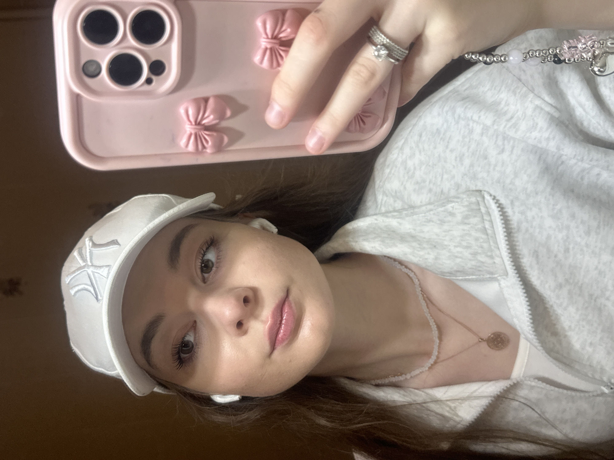 A young woman, appearing to be in her late teens or early twenties, is taking a selfie. She is wearing a white baseball cap with a logo, white earbuds, a light grey zip-up jacket over a white top, and a layered necklace with a circular pendant. She is also wearing rings on her right hand, which is holding a pink phone case with bow embellishments. Her expression is neutral, and she is looking towards the camera. The background appears to be an indoor setting with a wooden textured wall. There are no specific indicators in the image to suggest a location within the city of Kazan, Russia.