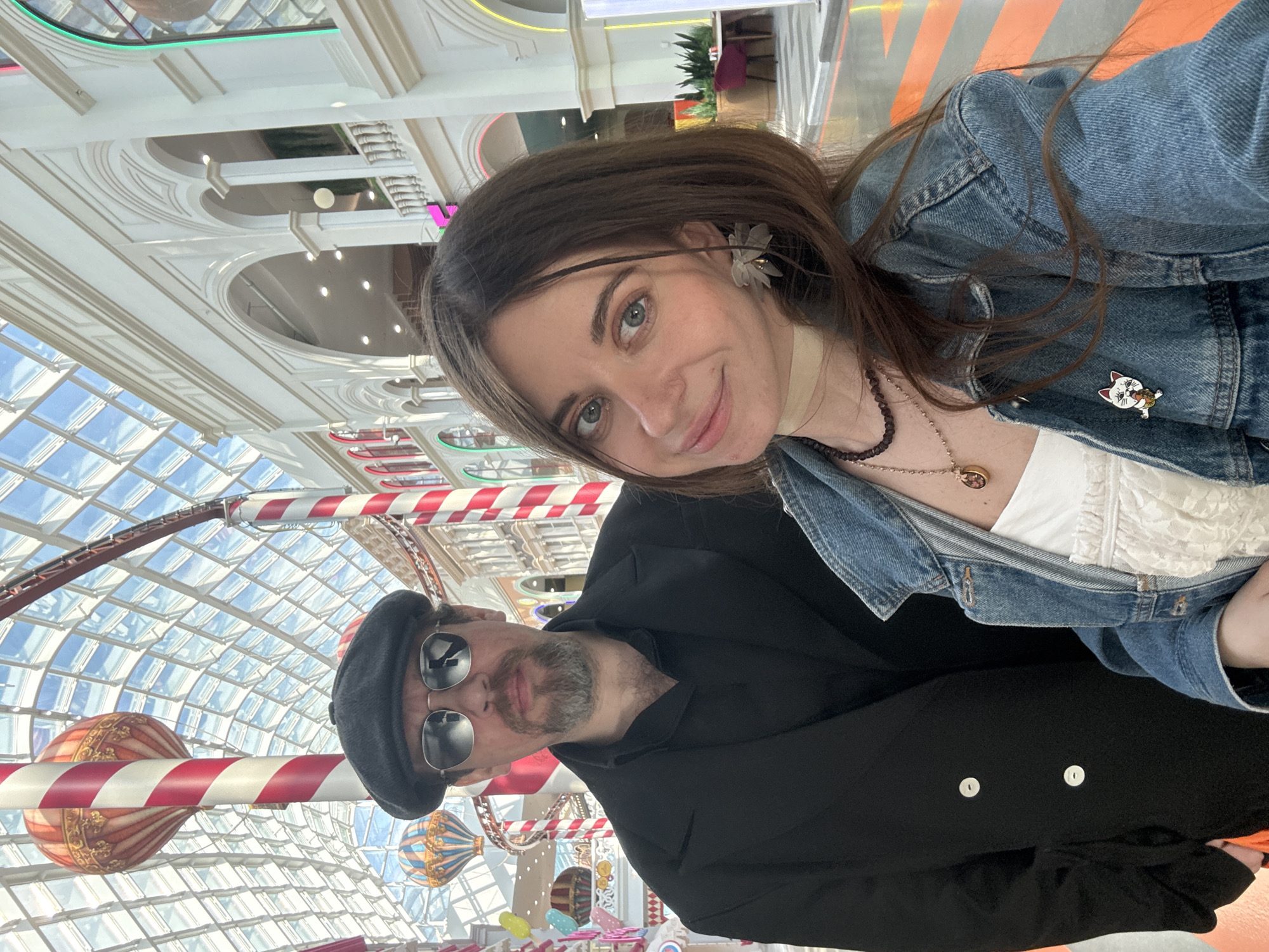 Two people are posing for a selfie in front of an interior that features a high ceiling with glass panels and decorative elements, including red and white striped poles and hot air balloon designs. The woman in the foreground is wearing a denim jacket over a white top, along with necklaces, and has a flower-shaped decoration in her hair. Next to her, a man is wearing sunglasses and a black hat. The location is likely a shopping mall or a similar indoor space. It is in GUM, a shopping center in Moscow, Russia.