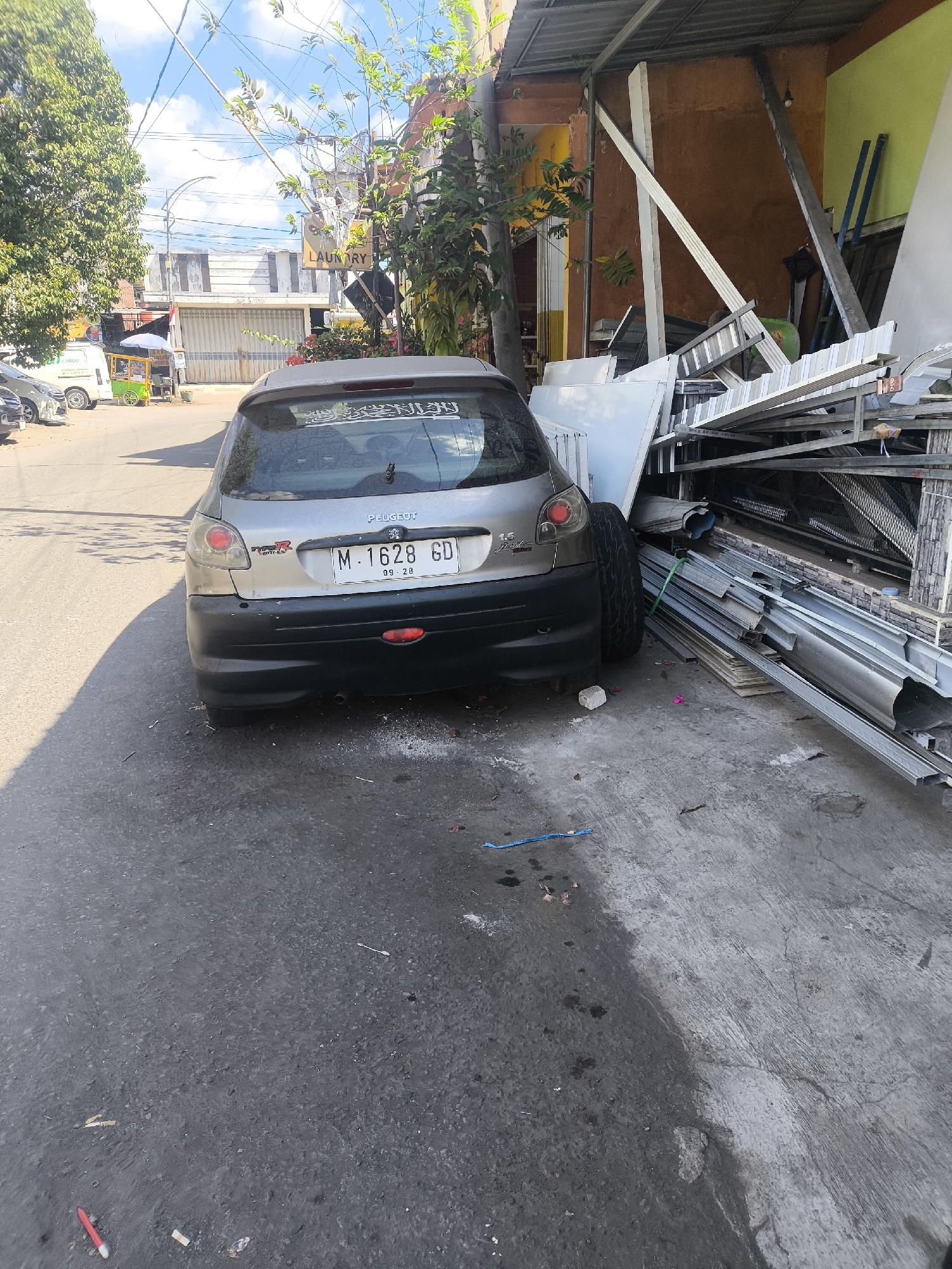 A silver Peugeot 206 hatchback is parked on the side of a paved street in Lumajang, Indonesia. The car's license plate reads M 1628 GD. Behind the car, to the right, is a cluttered area with various metal building materials, including long aluminum sheets and pipes, stacked haphazardly under a corrugated metal awning. A bright orange wall and a green wall are visible in the background. To the left of the car, a street lined with trees and buildings is visible, with a white van with "CARGO" written on it and a small vendor cart with a blue umbrella present. The sky is blue with scattered clouds. The ground is asphalt with some concrete areas. There are no people visible in the image.