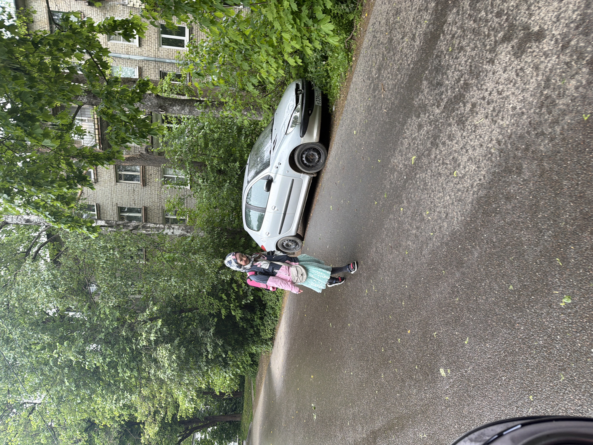 A girl wearing a headscarf, jacket, and dress stands on a wet asphalt road next to a silver car partially obscured by foliage on the right side of the road. A multi-story brick building is visible behind the foliage on the left side of the road. The scene appears to be in a residential area. The car has damage to its front end. Based on visual information, the location is Tokkari-Lend, Russia.