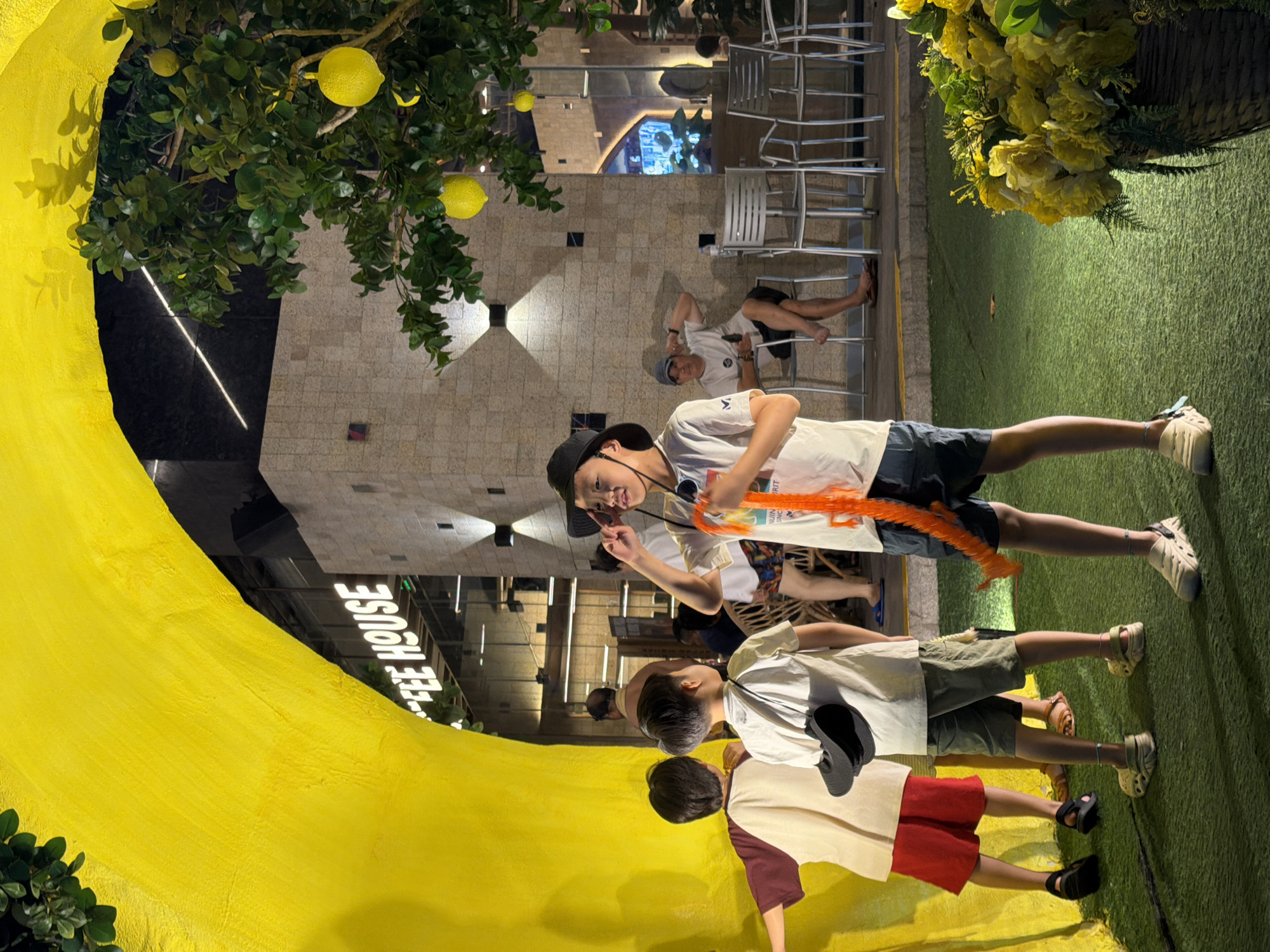 The image depicts an outdoor scene in Nha Trang Ward, Vietnam, likely at a cafe or restaurant with a prominent yellow architectural element reminiscent of a tunnel or archway. A cafe's sign, "COFFEE HOUSE," is visible in the background. In the foreground, several children are present. One boy, wearing a white t-shirt, black hat, and shorts, holds an orange toy. To his left, another boy in a white t-shirt and shorts stands facing away from the camera. A third child, to the left of the frame, wears a maroon and cream shirt and red shorts.
In the middle ground, a man wearing a grey cap and white t-shirt sits on a chair, with his legs crossed and one arm resting on his knee. Behind him, several metal chairs are arranged at tables. A tree with artificial lemons hangs with bright yellow fruits. The ground is covered in green artificial turf, and a basket filled with yellow flowers sits to the right of the frame. The lighting suggests it is evening.